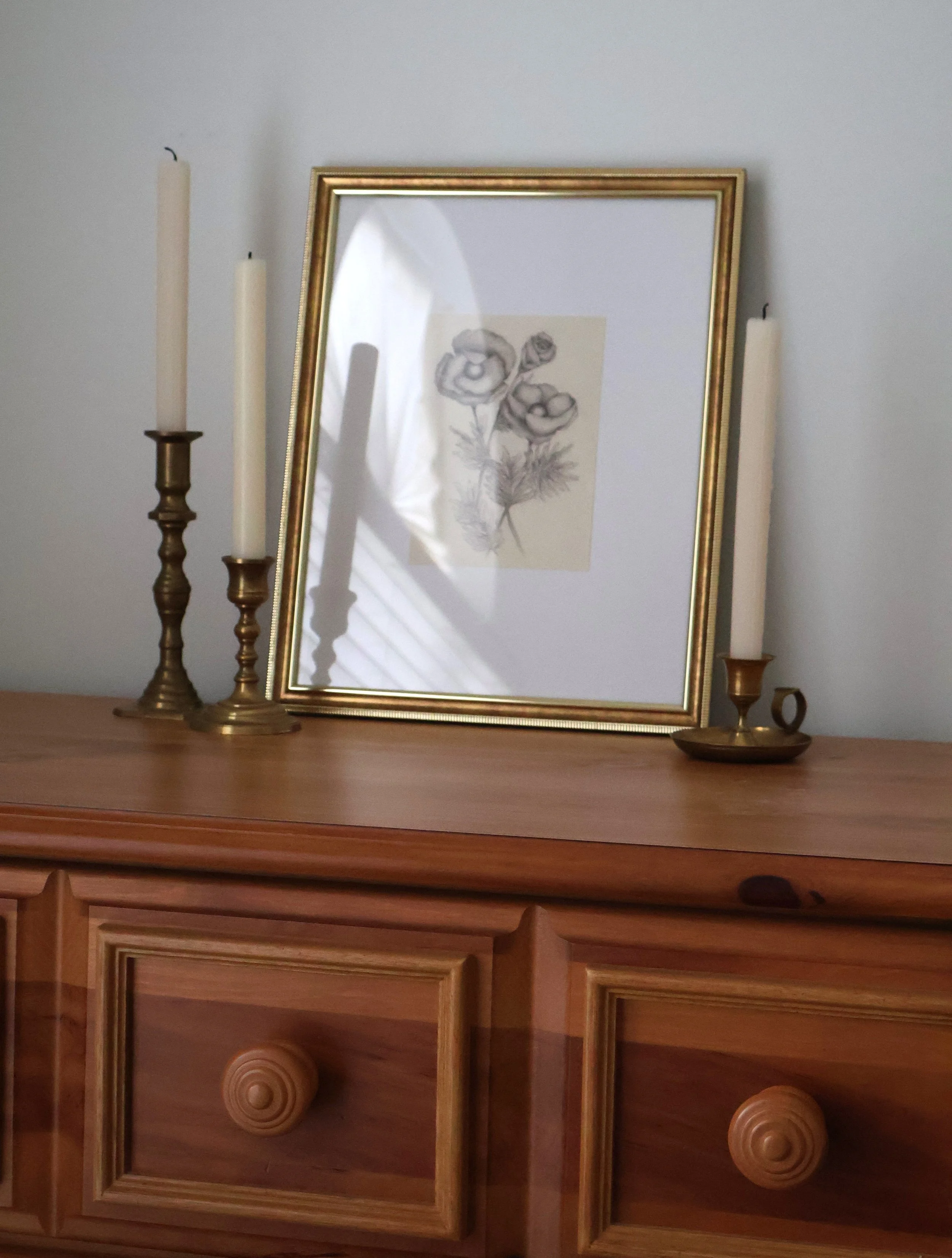 A wooden dresser with a gold-framed mirror, three candlesticks with unlit candles, and a framed floral drawing in the reflection.