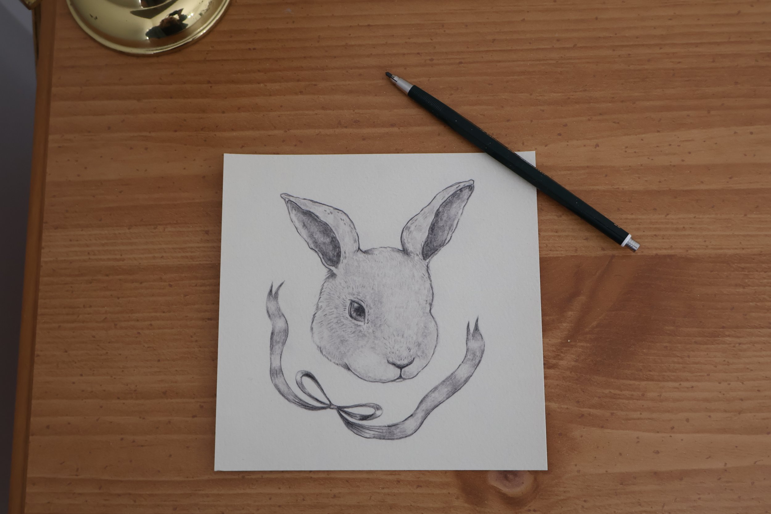 A pencil sketch of a rabbit's face with a ribbon around its neck, placed on a white sheet of paper on a wooden table.