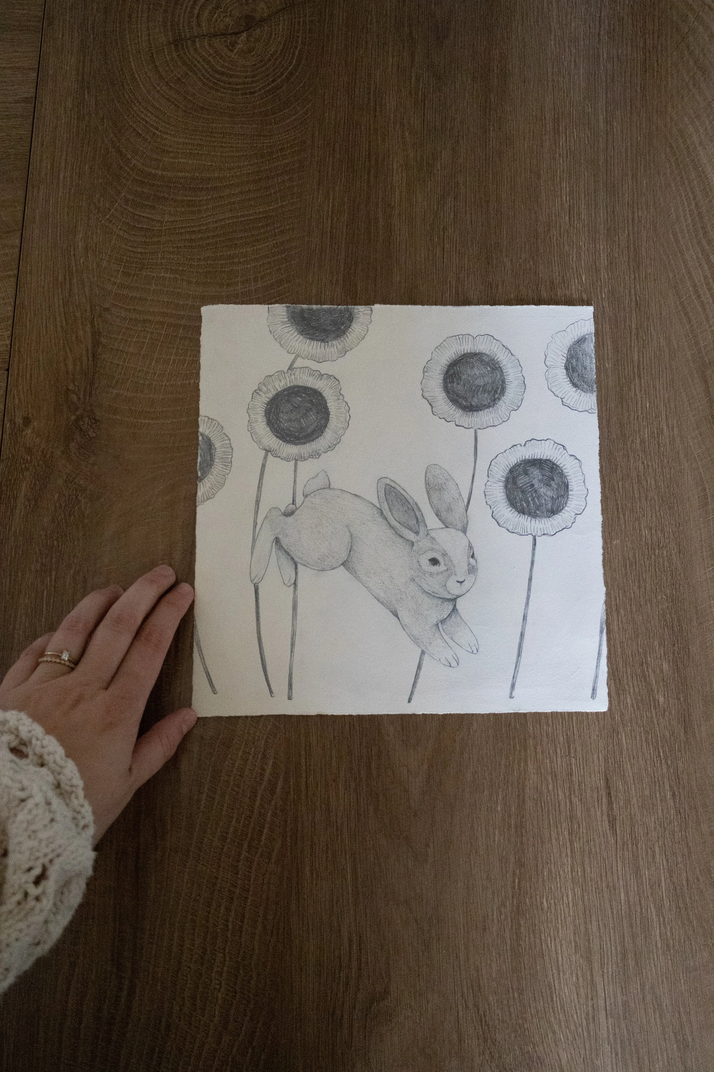 A hand holding a black and white drawing of a rabbit surrounded by large sunflowers on a wooden surface.