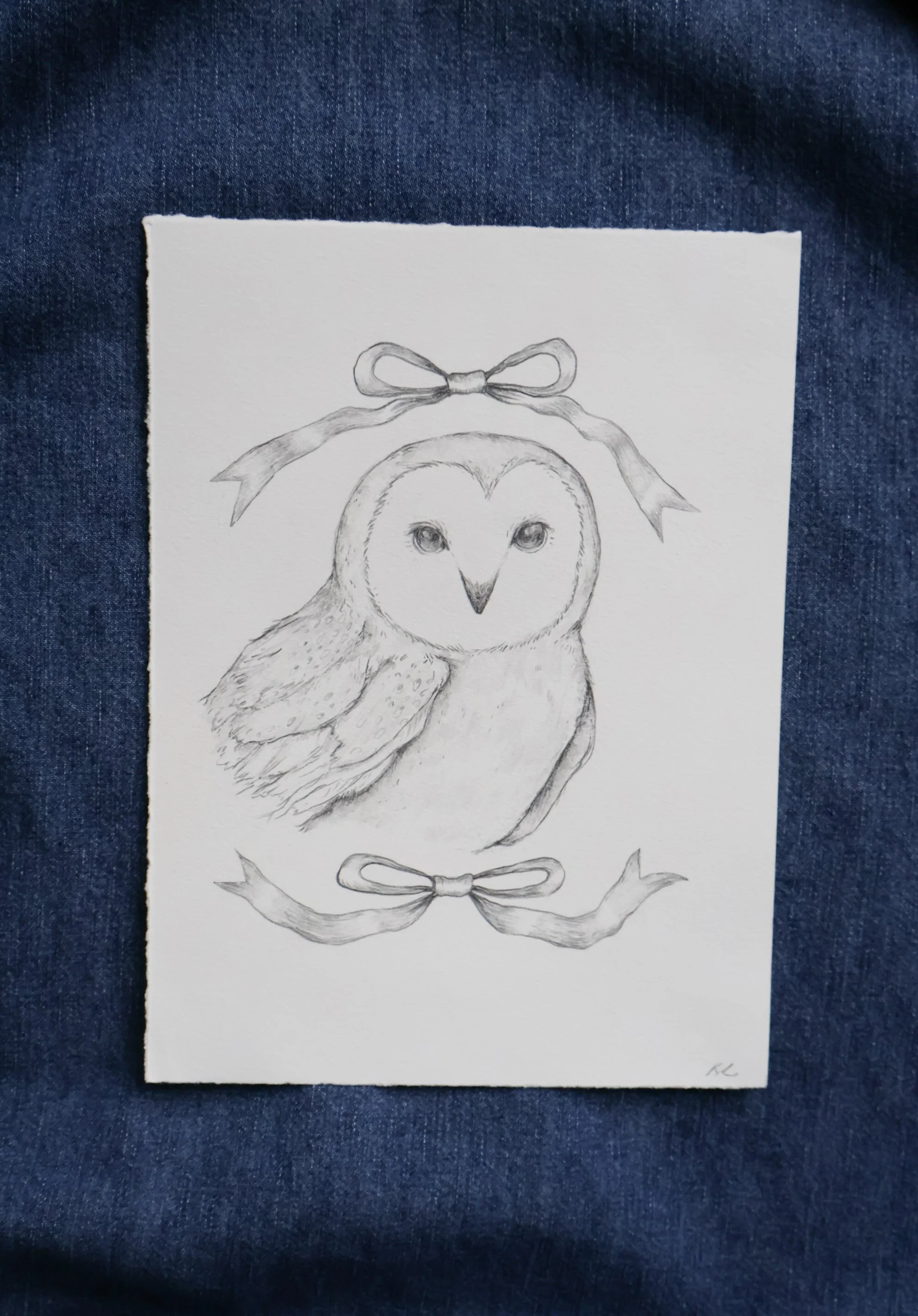 Pencil sketch of an owl with bows at top and bottom, on white paper, placed on denim fabric.