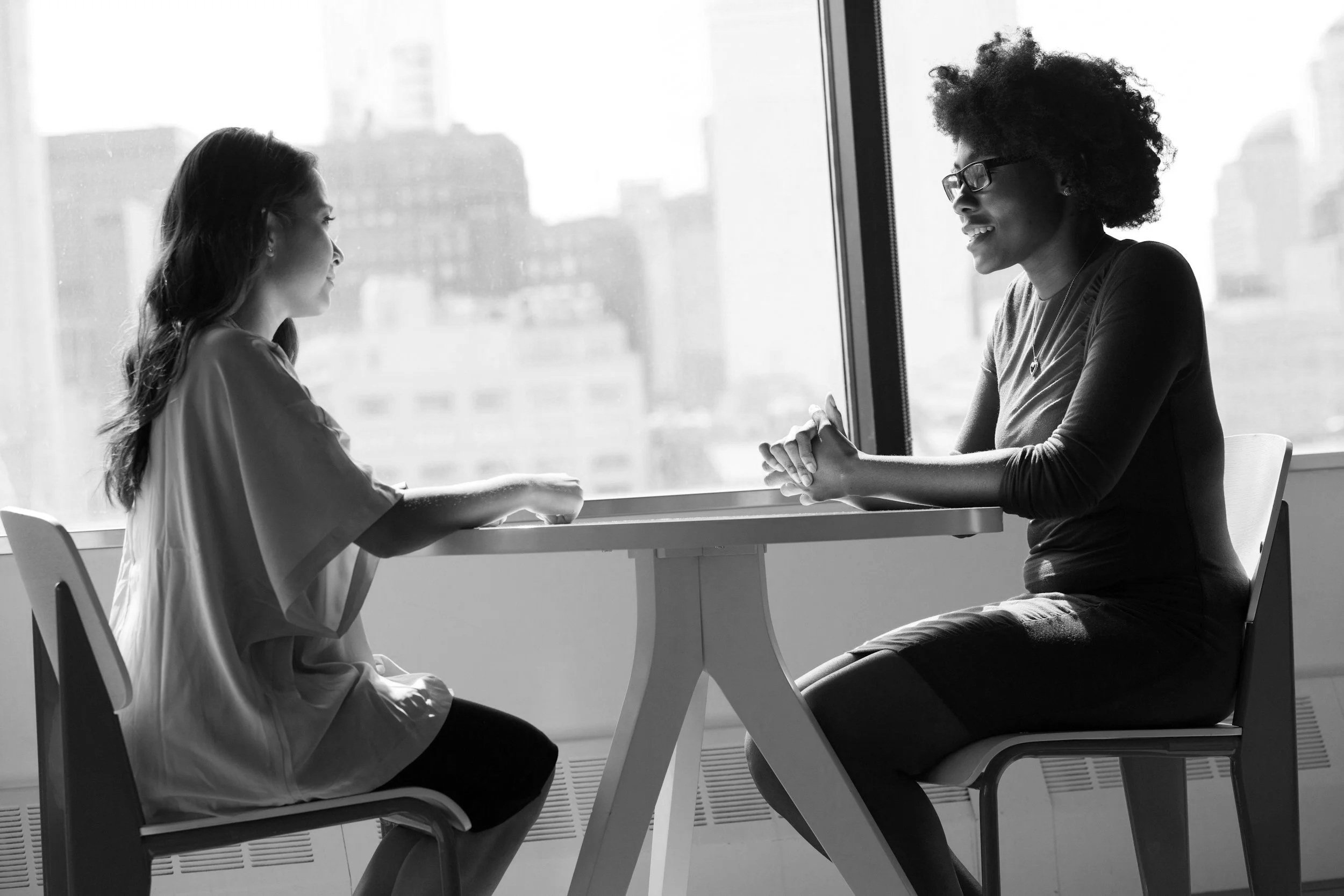 Two women sit across from each other at a table in front of a large window, engaging in conversation. One woman is wearing a loose blouse, and the other has curly hair and glasses.