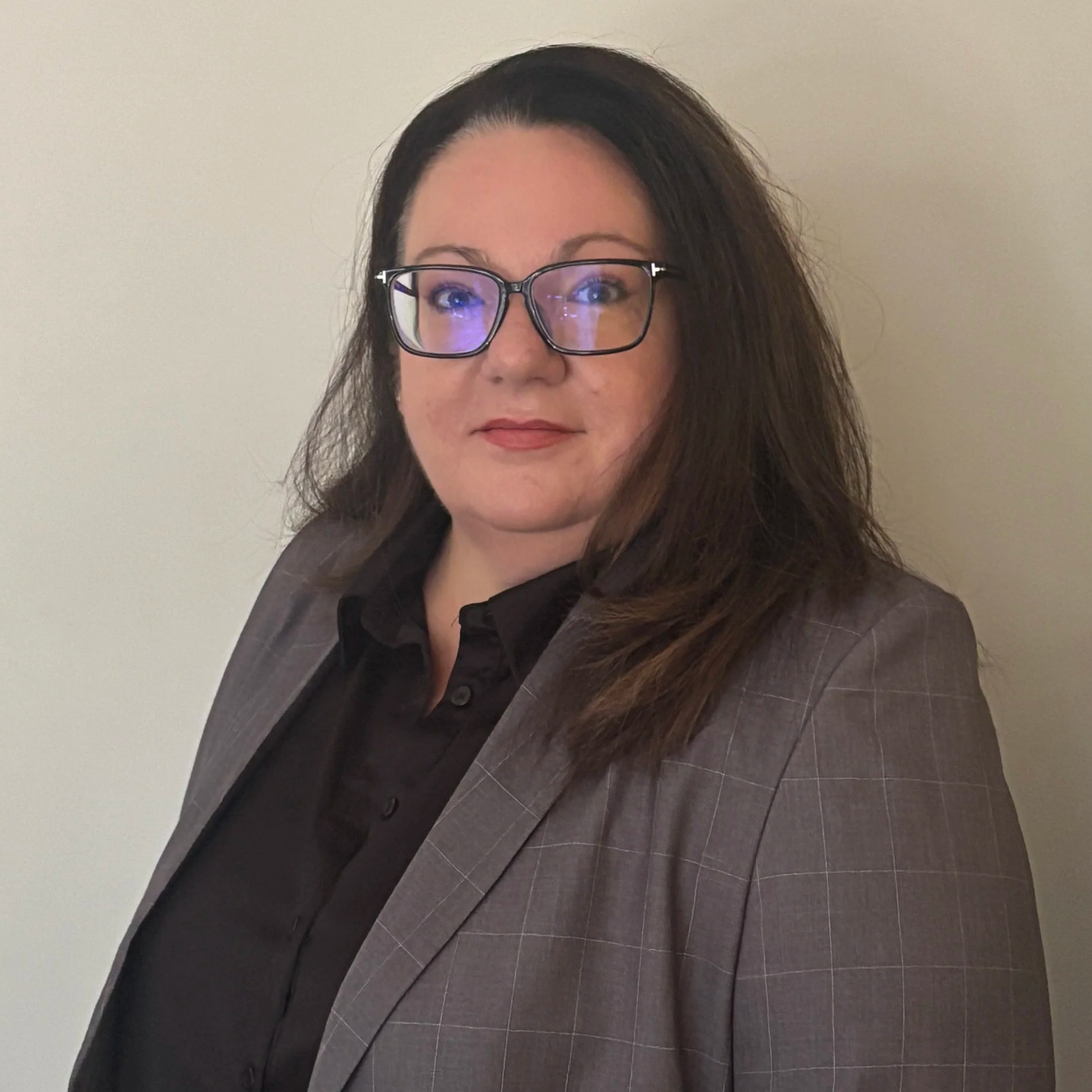 A woman with glasses, dark brown hair, wearing a gray checkered blazer and a black shirt, standing against a plain wall.