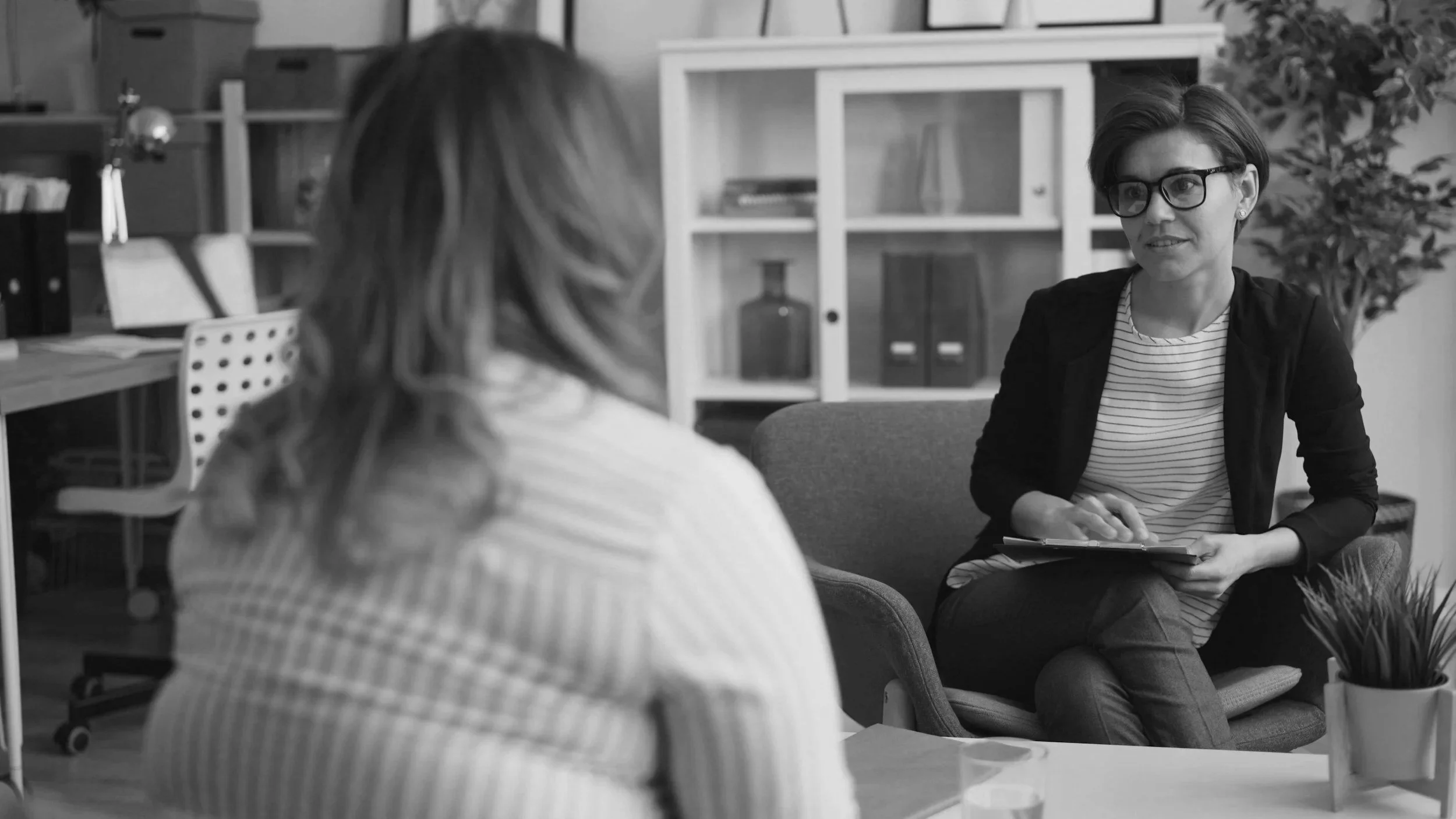 Two women having a conversation in an office setting, one with glasses and short hair, holding a notebook, and the other with long hair, seen from behind.
