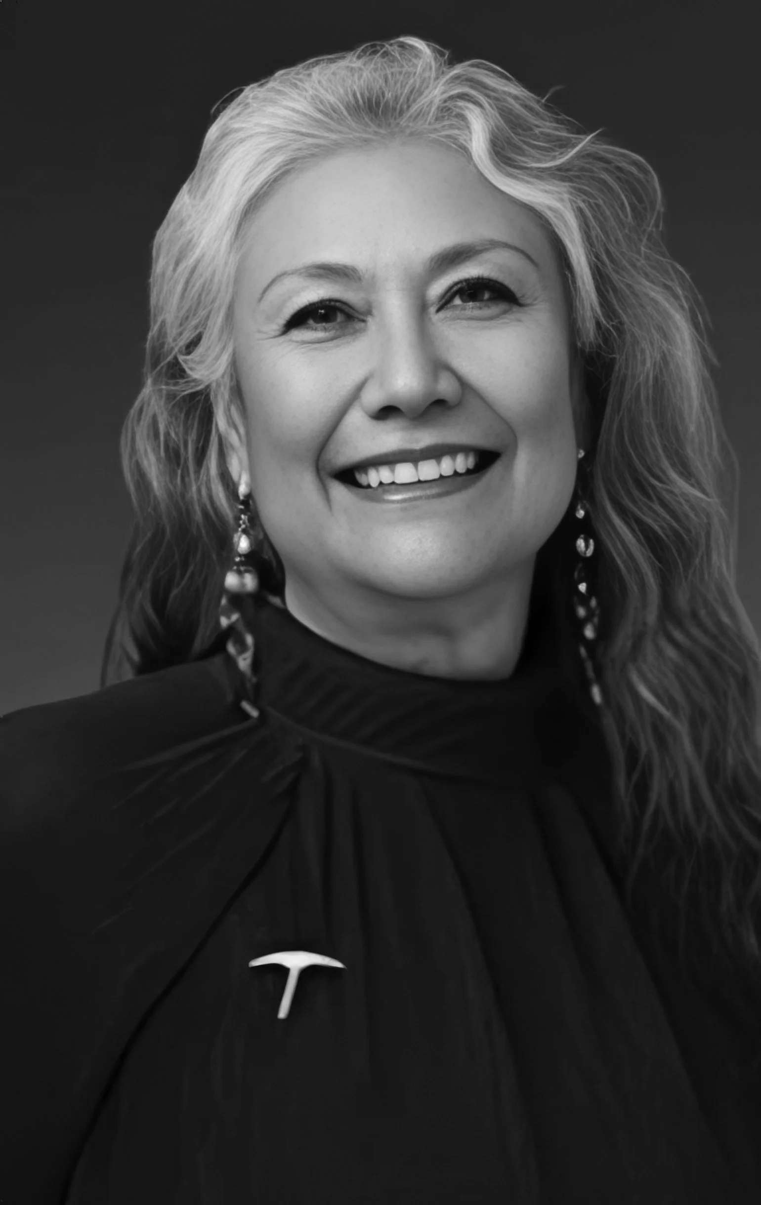Black and white portrait of a smiling woman with long, wavy hair wearing earrings and a dark top, against a dark background.