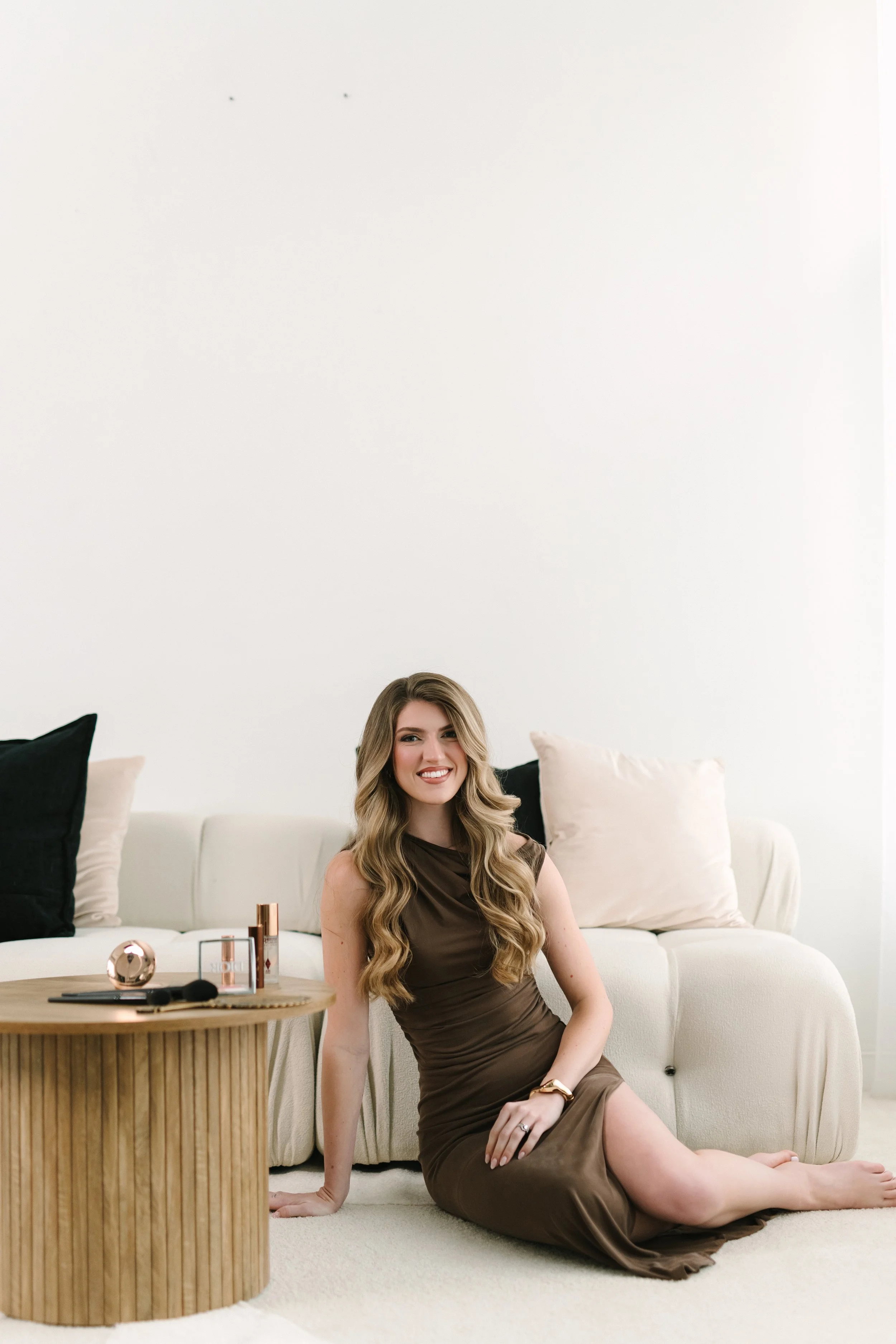 Woman with long wavy hair wearing a brown dress, seated on the floor beside a cream couch with black and beige pillows, smiling at the camera with a small wooden table near her holding makeup products and a mirror.