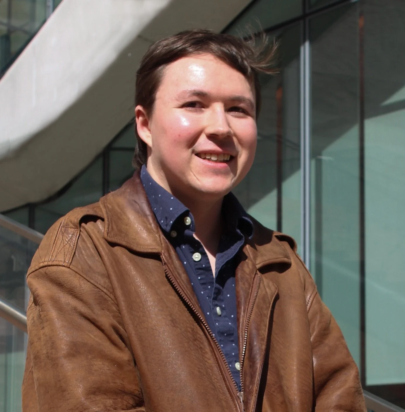 A young man with dark hair smiling outdoors in front of a modern building with glass windows and a concrete staircase, wearing a brown leather jacket and a navy blue shirt with white polka dots.