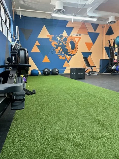 Interior of a gym with artificial turf, gym equipment, medicine balls, and a colorful wall mural with a person lifting weights
