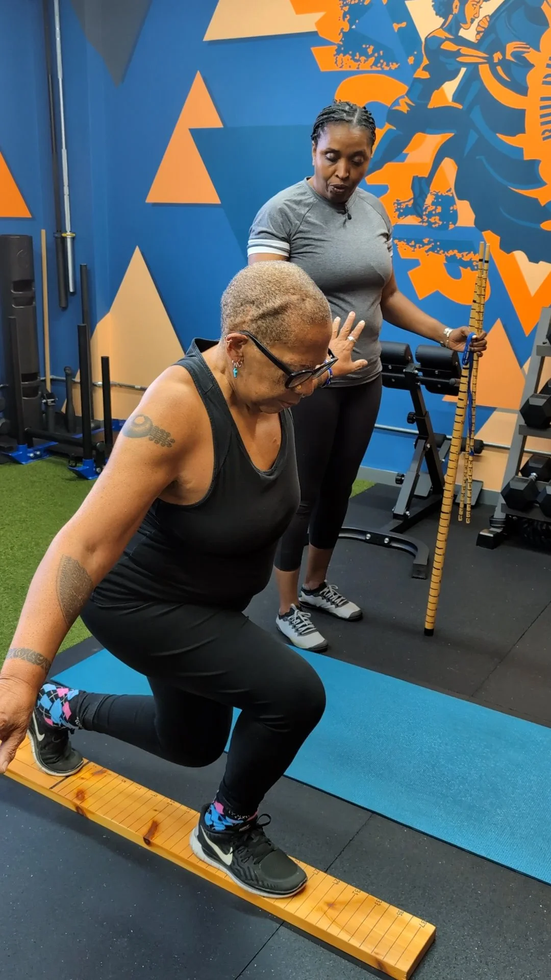 Two women exercising in a gym, one is balancing on a wooden balance beam while the other observes, with gym equipment and blue and orange wall art in the background.