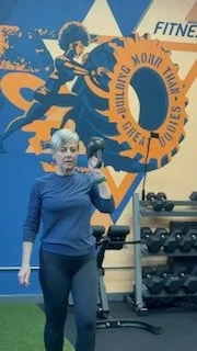 An elderly woman in a blue long-sleeve shirt standing in a gym, holding a large, orange ring with the words 'Youth, Moving, Learning' on it. There are weights and gym equipment behind her.