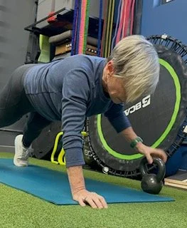 Elderly woman performing a kettlebell workout on a blue exercise mat in a gym.