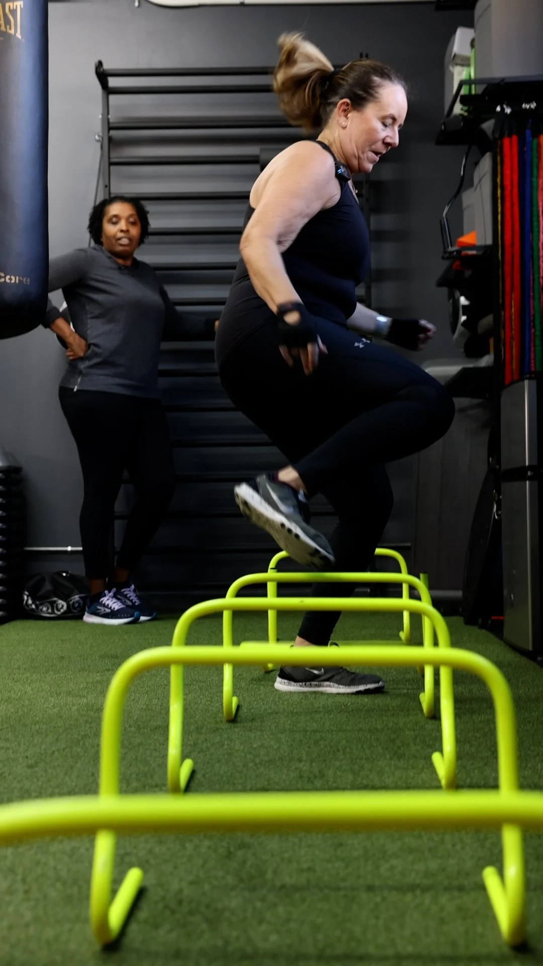 A woman performing box jumps over yellow hurdles in a gym, with another woman watching in the background.