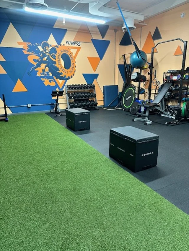 Fitness gym with black rubber flooring and green turf area, featuring workout equipment such as dumbbells, a plyometric box, ropes with a medicine ball, and a wall with a running graphic and colorful triangles.
