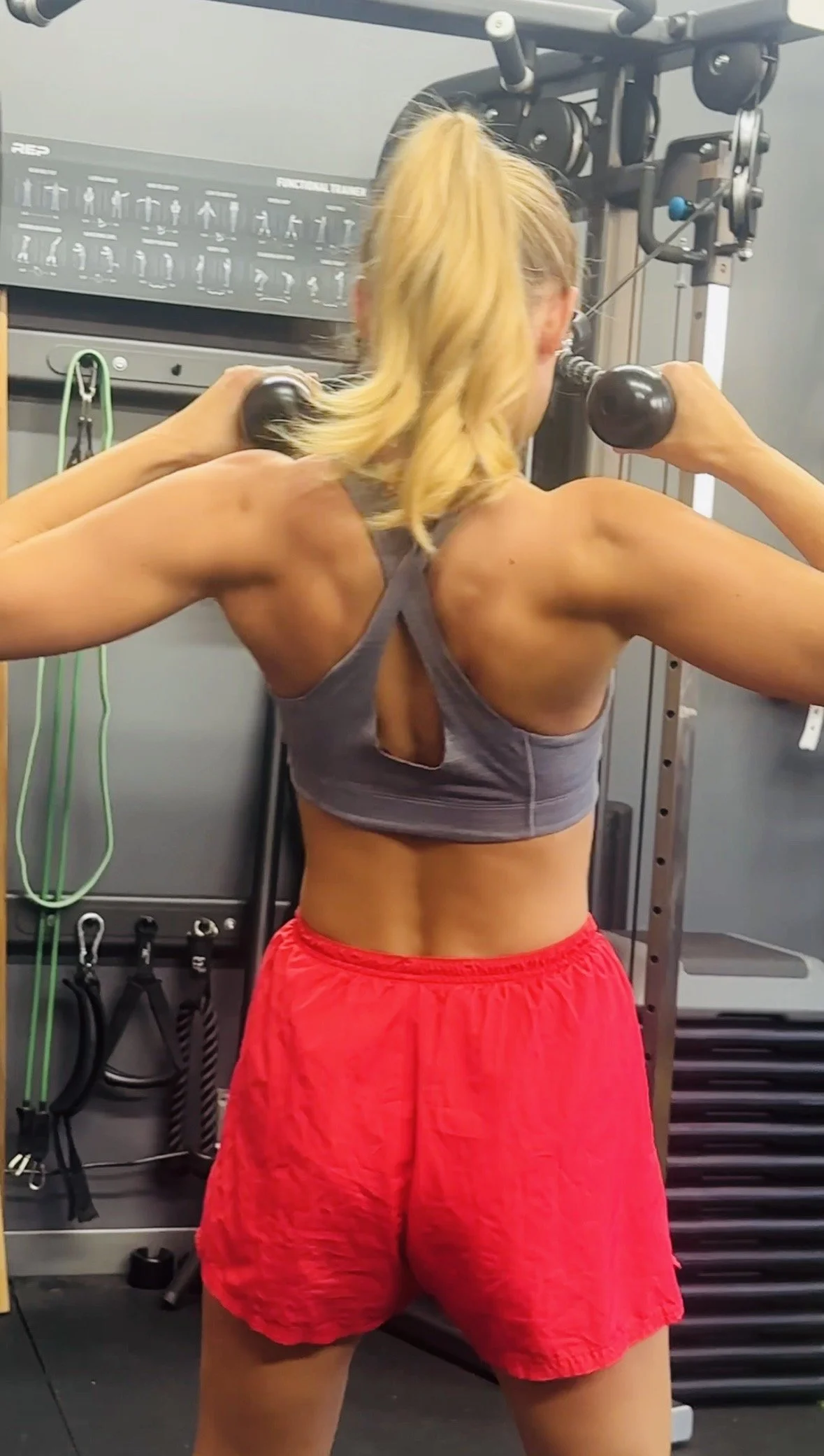 Woman with blonde hair in a ponytail doing a shoulder press exercise with dumbbells in a gym, facing away from the camera.