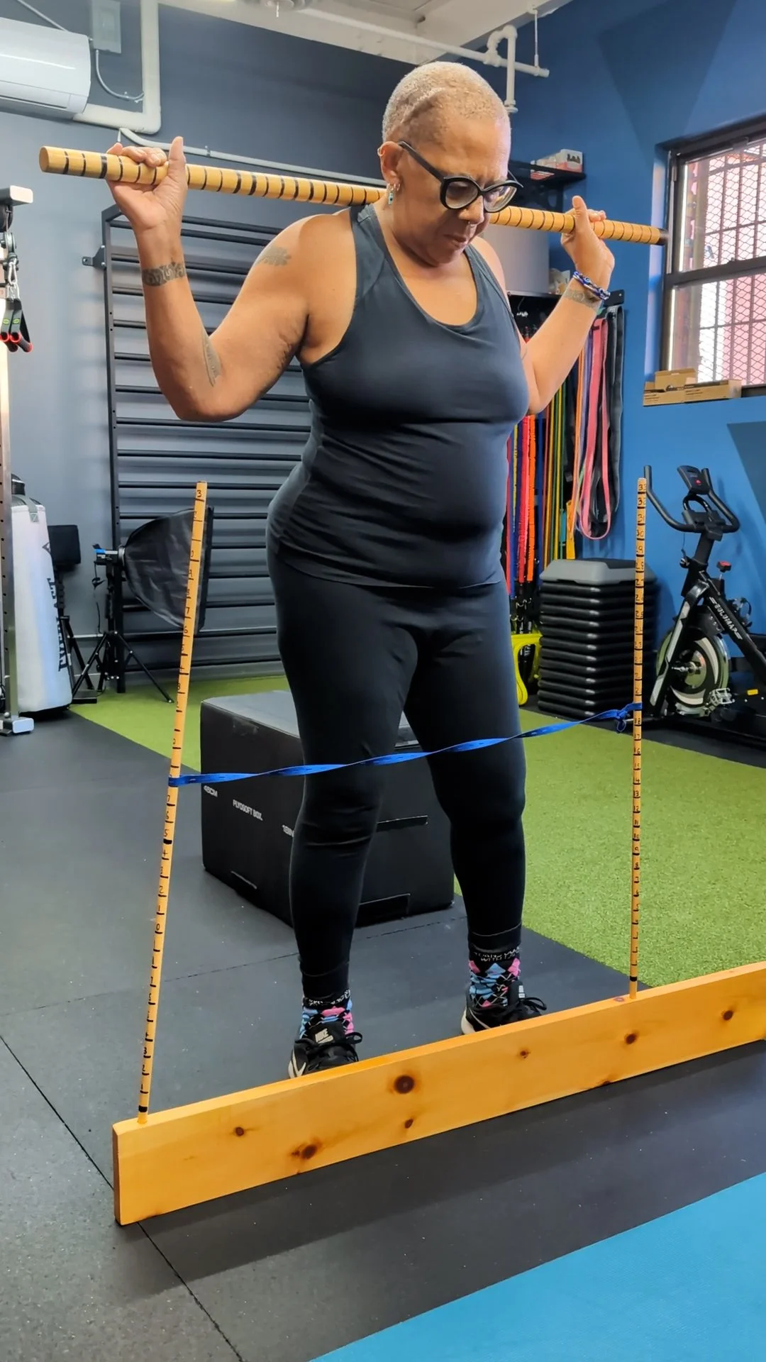 Woman doing squat exercise with a wooden frame and measuring markings, in a gym with fitness equipment and resistance bands.