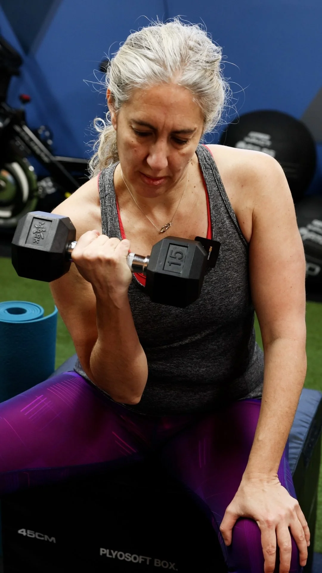 An older woman with gray hair exercising in a gym, lifting a 15-pound dumbbell in her right hand, while seated on a purple mat. She is wearing a gray tank top with red straps and purple leggings.
