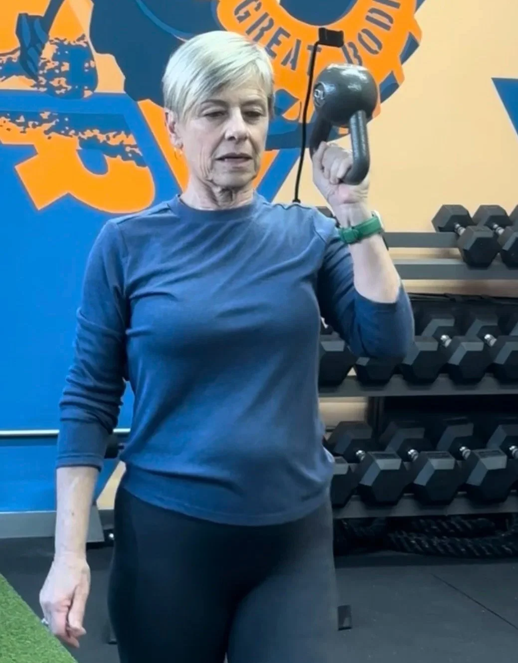 A woman with short blonde hair wearing a navy long-sleeve shirt and black workout pants, holding a weight plate in a gym.