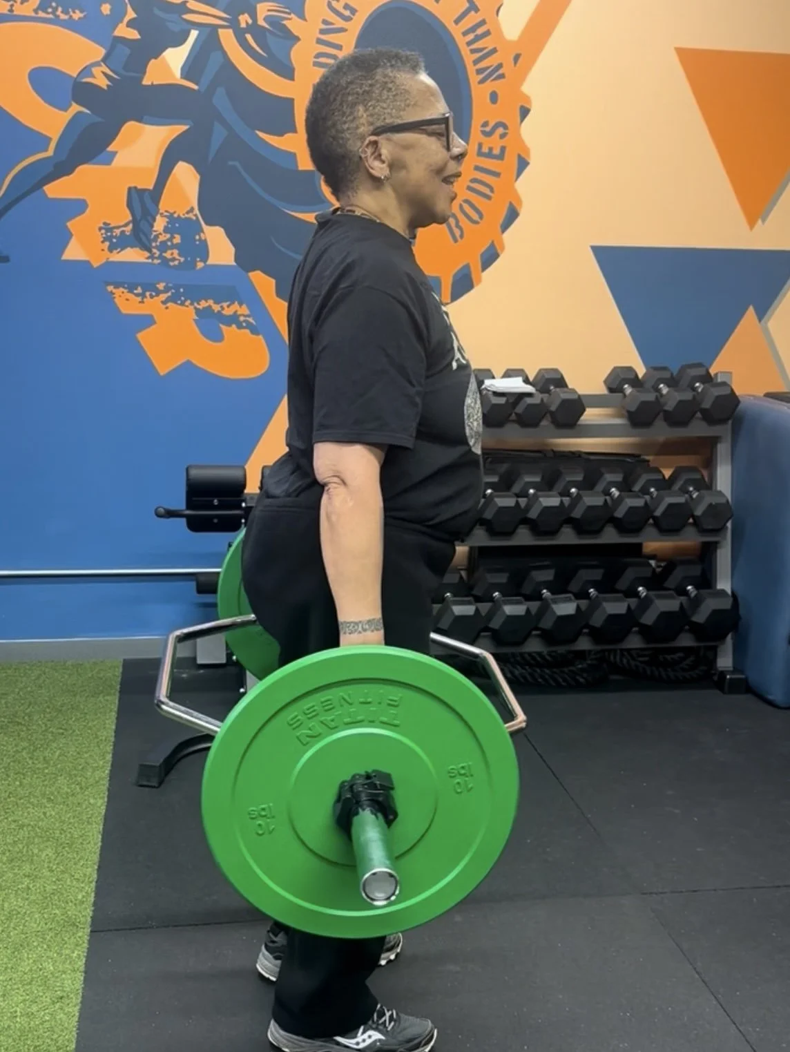 Woman lifting a barbell with a green weight plate in a gym with a blue and orange wall featuring a logo and a rack of dumbbells.