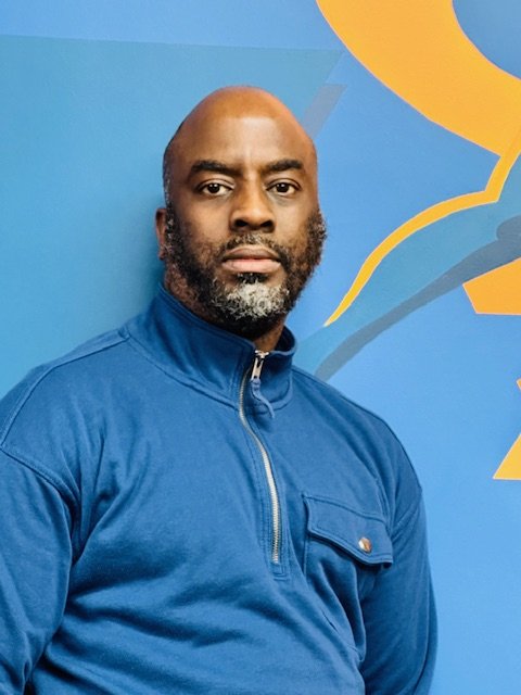A man with a bald head and a beard wearing a blue zip-up jacket standing in front of a blue wall with yellow and dark gray abstract shapes.