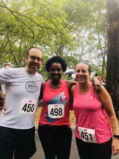 Three smiling women stand together outdoors among trees, wearing athletic clothing and race bibs, indicating participation in a running event.