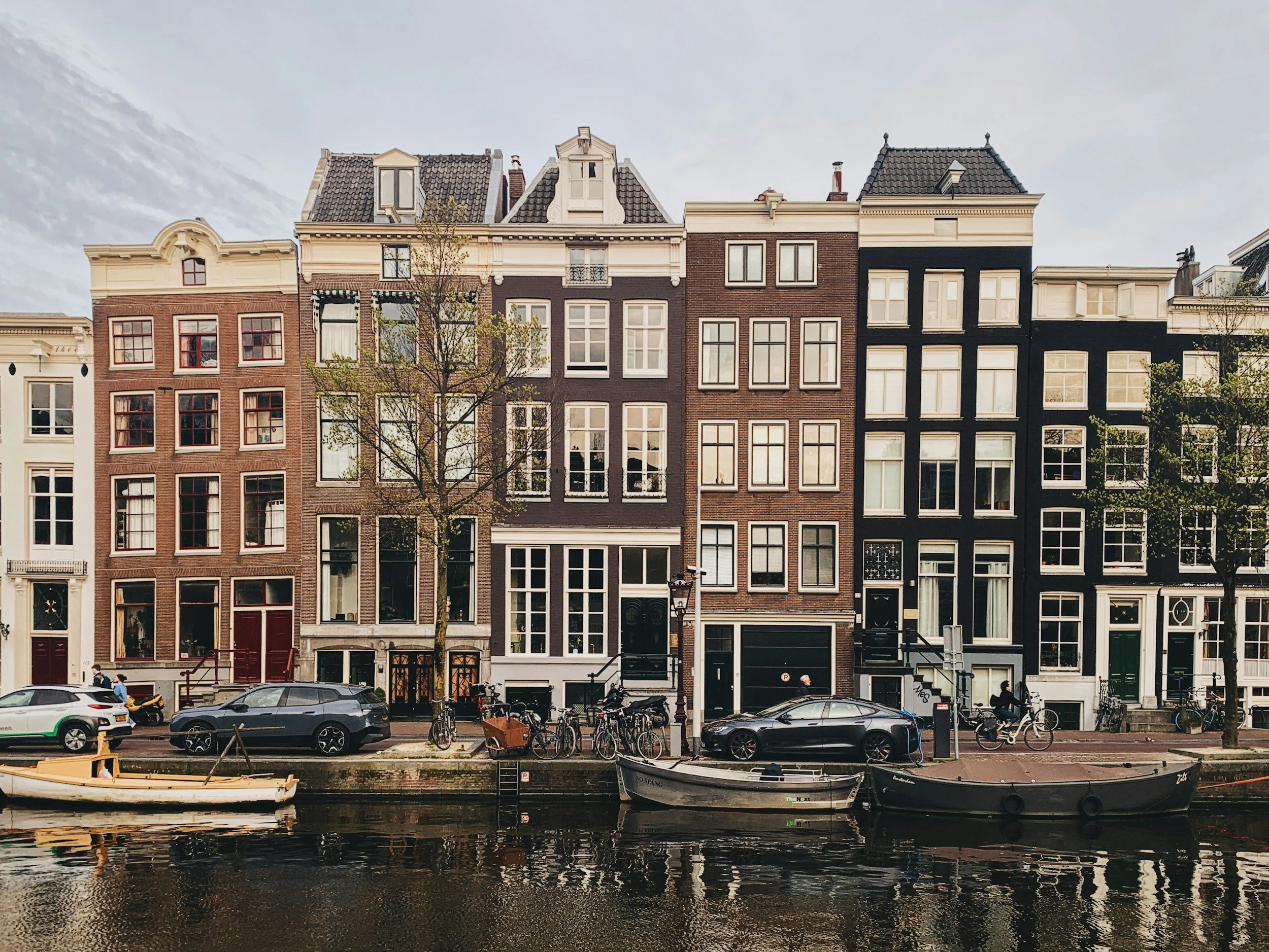 A row of narrow European-style townhouses along a canal, with boats docked in the water and parked cars on the street in front.