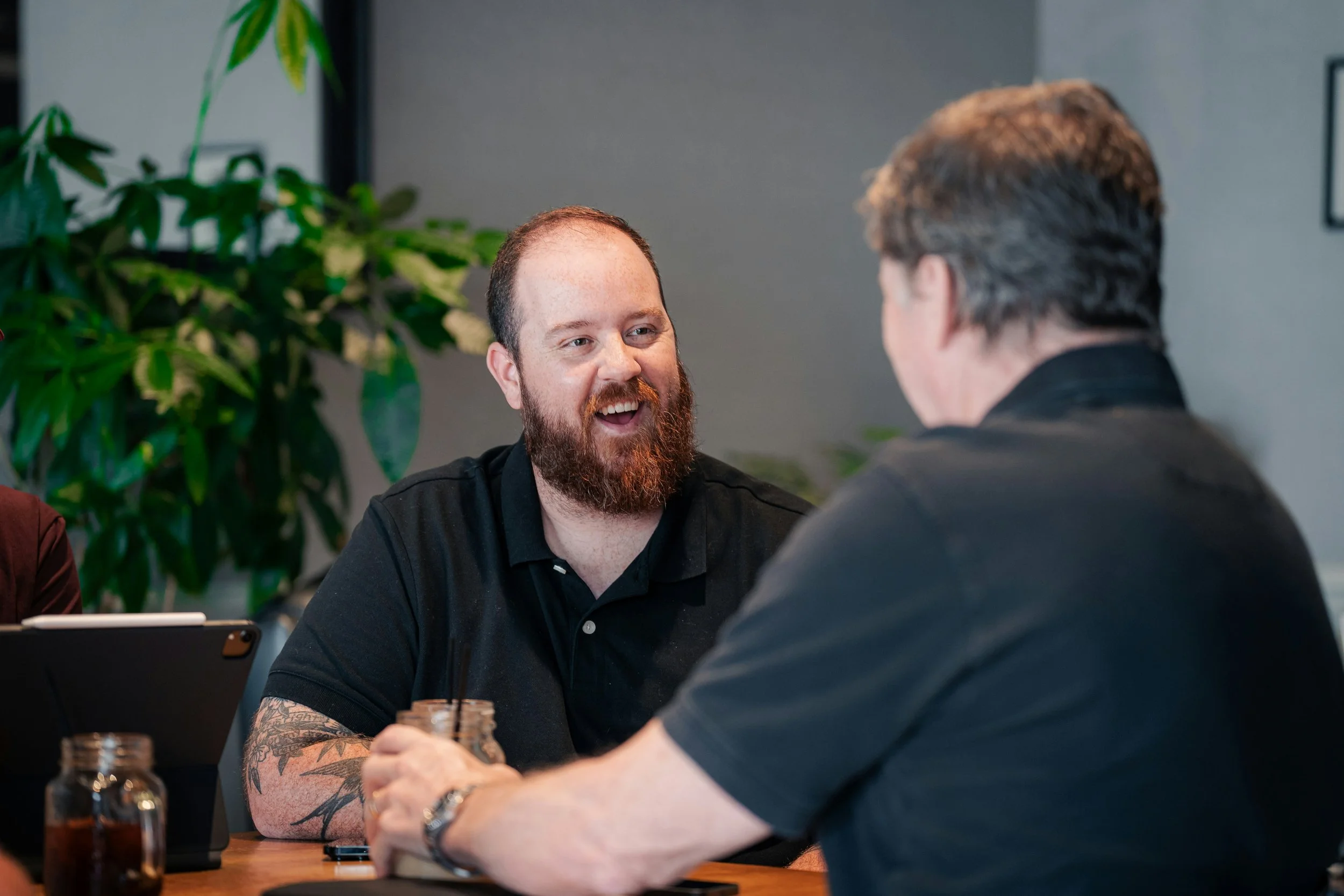 Two men having a conversation at a table with drinks and a tablet, with green plants in the background.