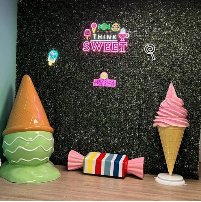 Decorative ice cream sculptures and neon signs on a black glittery wall. An ice cream cone sculpture on the left, a wrapped candy in the middle, and a soft serve ice cream cone sculpture on the right. Neon signs read "THINK SWEET" and "LIFE IS SHORT, MAKE IT SWEET."