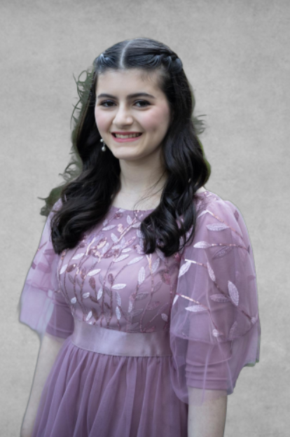 A young woman with dark, wavy hair wearing a purple dress with floral embroidery and puffed sleeves, smiling against a plain wall background.