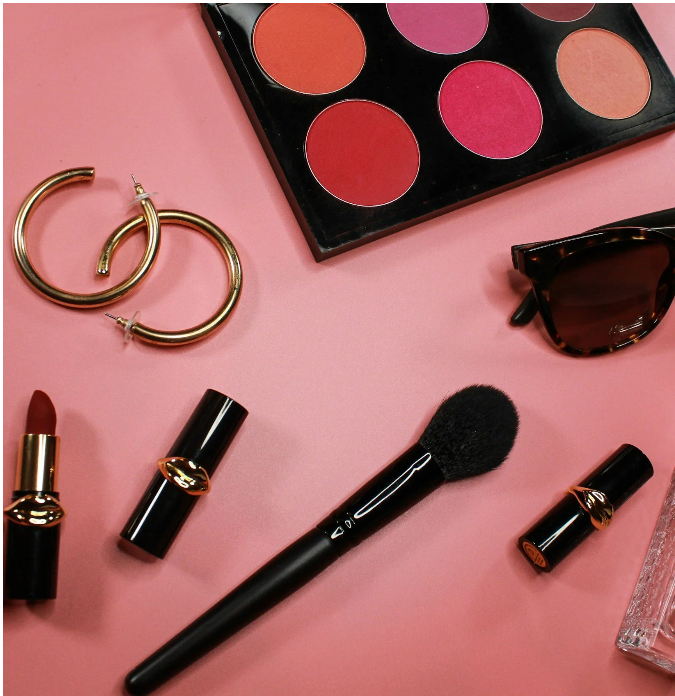 Makeup brush, lipsticks, eyeshadow palette with pink and red shades, gold hoop earrings, tortoiseshell sunglasses, and a perfume bottle on a pink background.