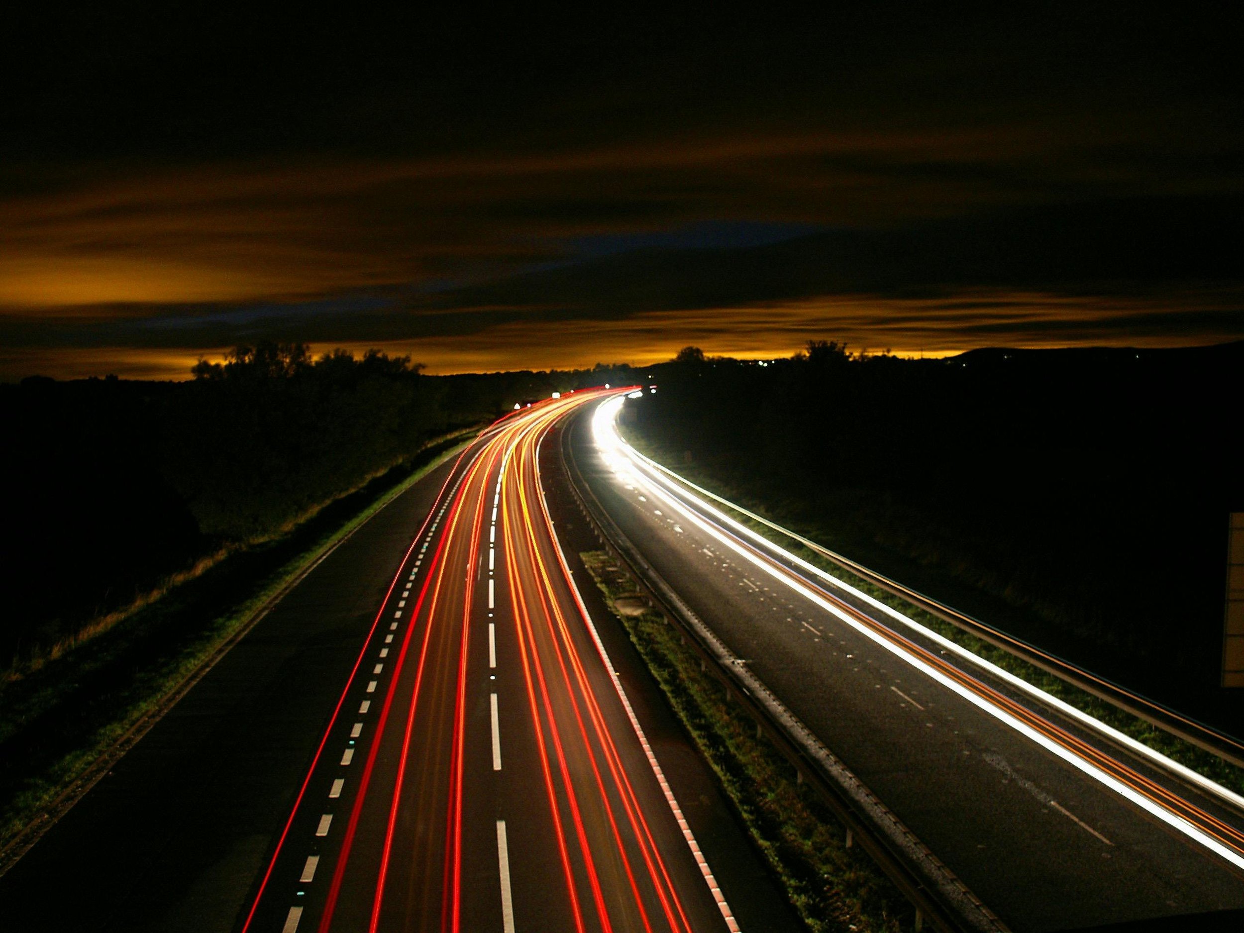 Long exposure photo of a highway at night with streaks of red and white lights from moving vehicles, dark sky with clouds and a faint orange glow near the horizon.
