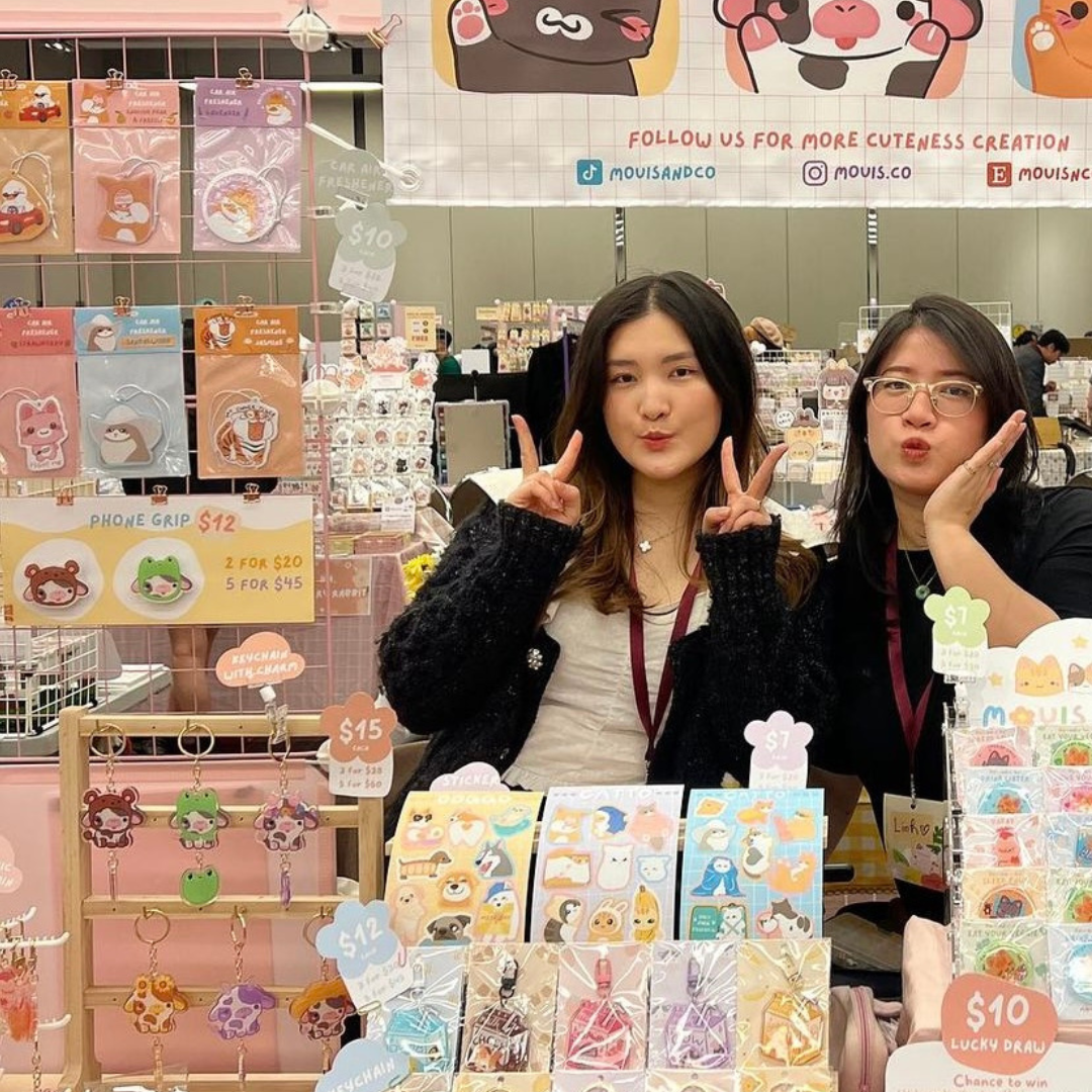 Two women standing behind a table of cute stickers and accessories at a craft fair or market.