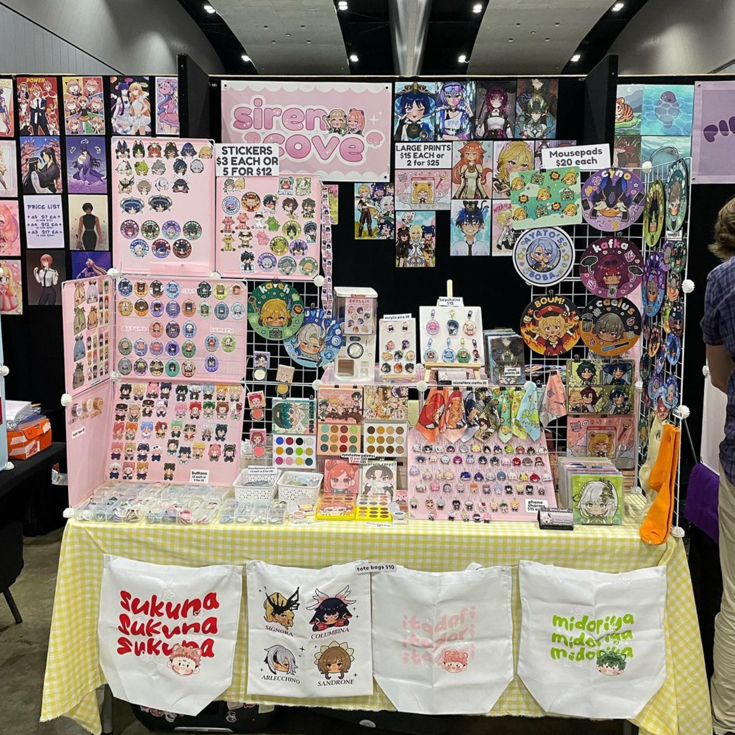 A vendor's booth at an anime and kawaii merchandise convention, featuring stickers, buttons, keychains, badges, and art prints with cute and anime-themed designs, displayed on grids and boards, with signs indicating prices and special offers.