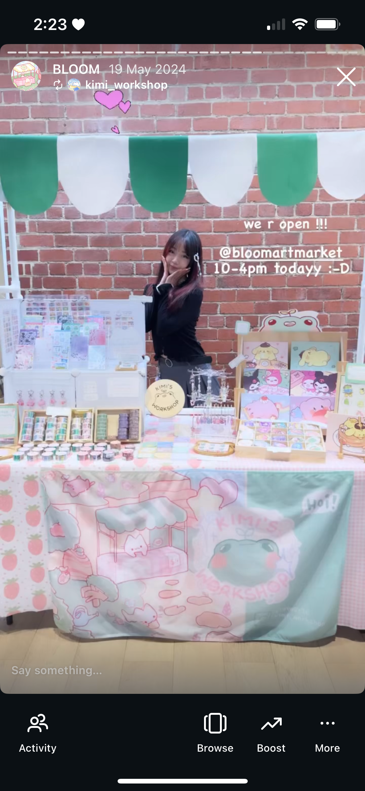 A woman standing behind a market stall with colorful stationery, stickers, and art prints, decorated with a pink and pastel-themed tablecloth and a green and white banner overhead, against a brick wall.