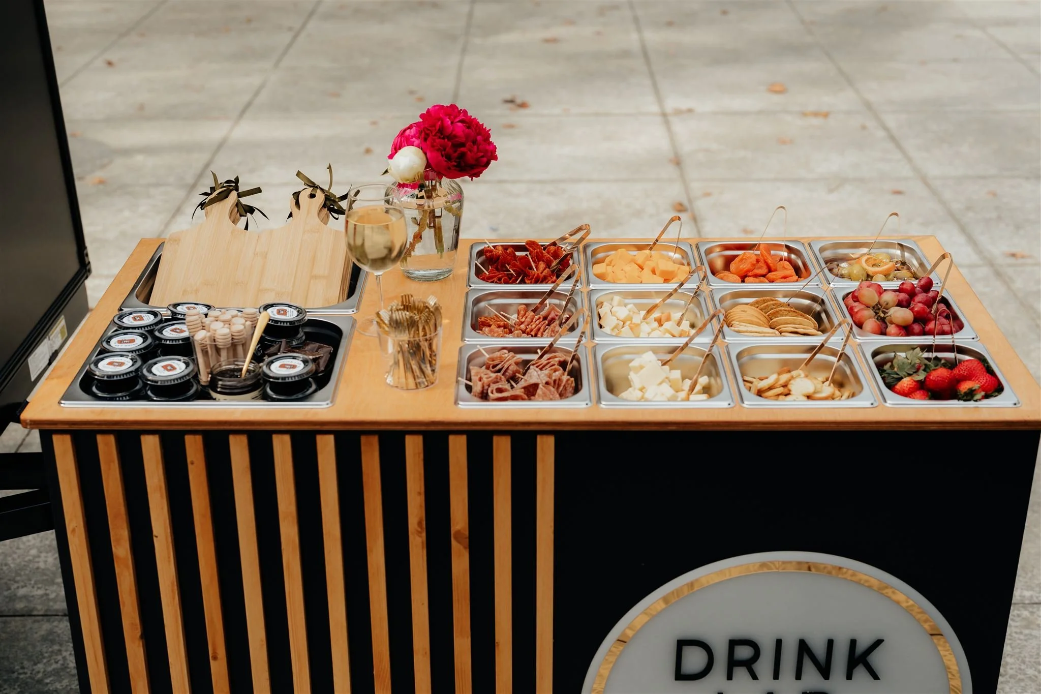 A small wooden cart with a variety of fresh fruit, cheese, and snacks, including strawberries, grapes, cheese cubes, dried fruit, crackers, and small containers of jam. There is a glass of champagne, a small glass jar of toothpicks, and a flower arrangement with pink flowers in a clear vase, on the cart. The background is a paved outdoor area.