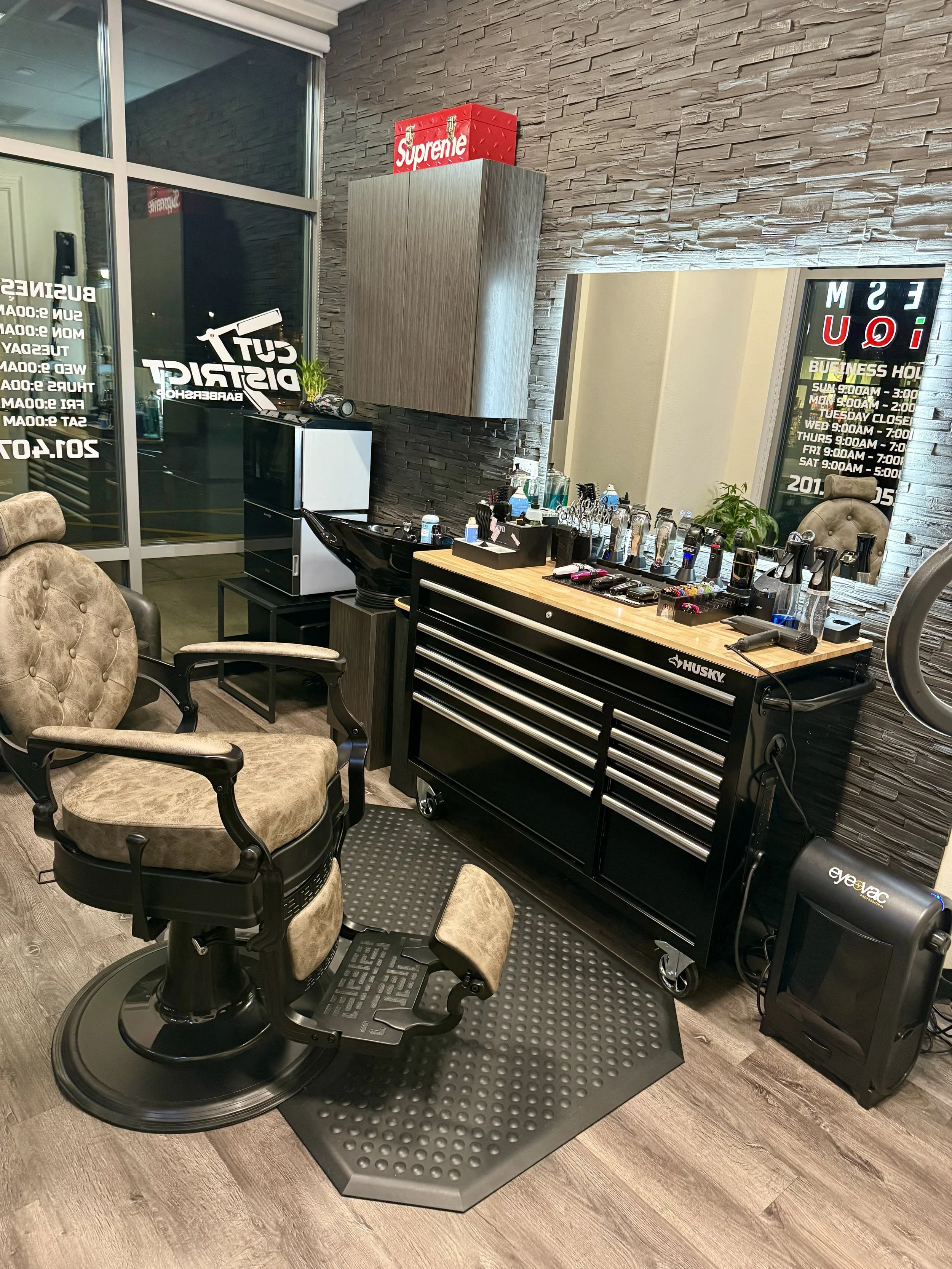 Barber’s station with vintage-style barber chair, various hair styling tools, and products, in a modern salon with a brick accent wall and a window with shop hours