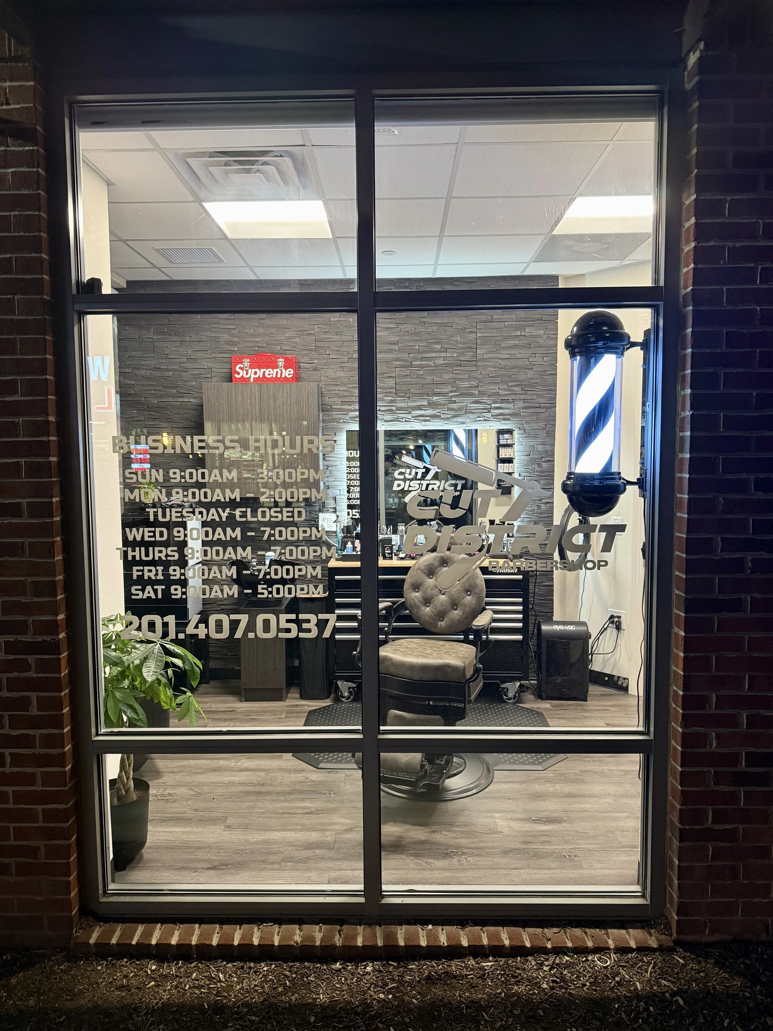 View of a barber shop through a glass window. The shop has a barber chair, a plant, a wall-mounted mirror, and a barber pole. The shop's business hours are written on the window.