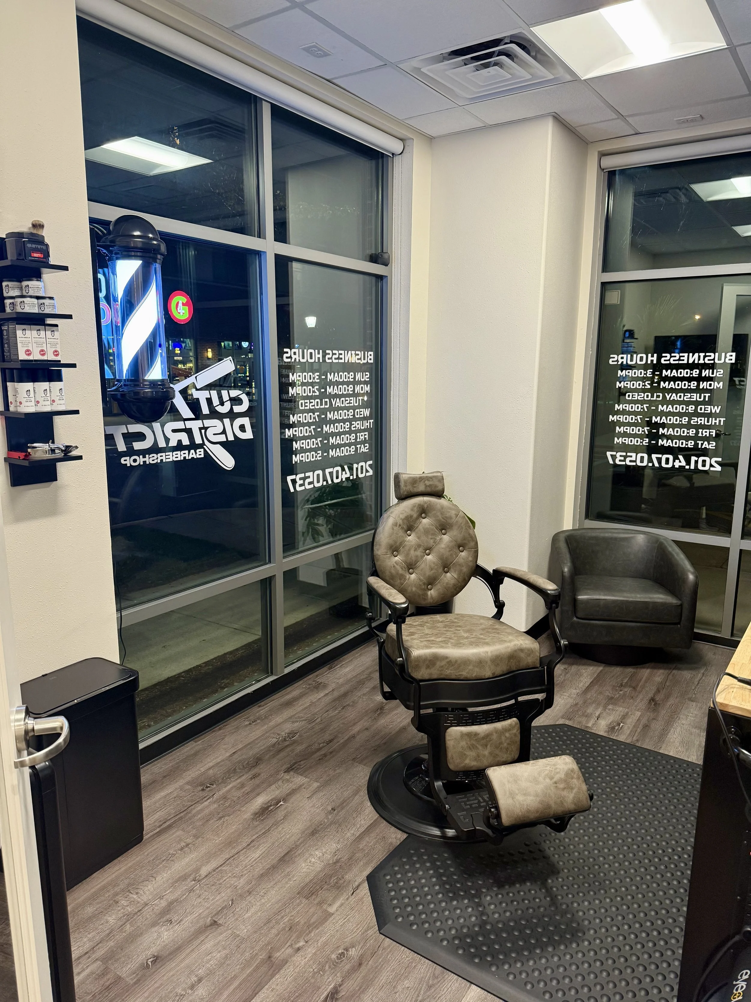 Barbershop interior with a vintage barber chair, a black armchair, large windows with signs listing services and contact information, and a small shelf with barber supplies.