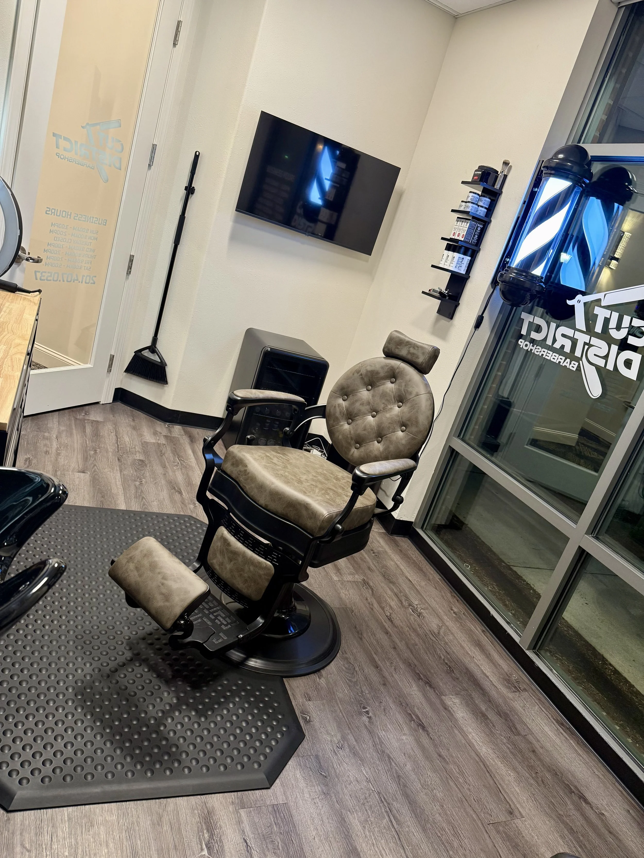 Barbershop interior with vintage barber chair, large window with stickers, wall-mounted TV, broom, and shelving with hair products.