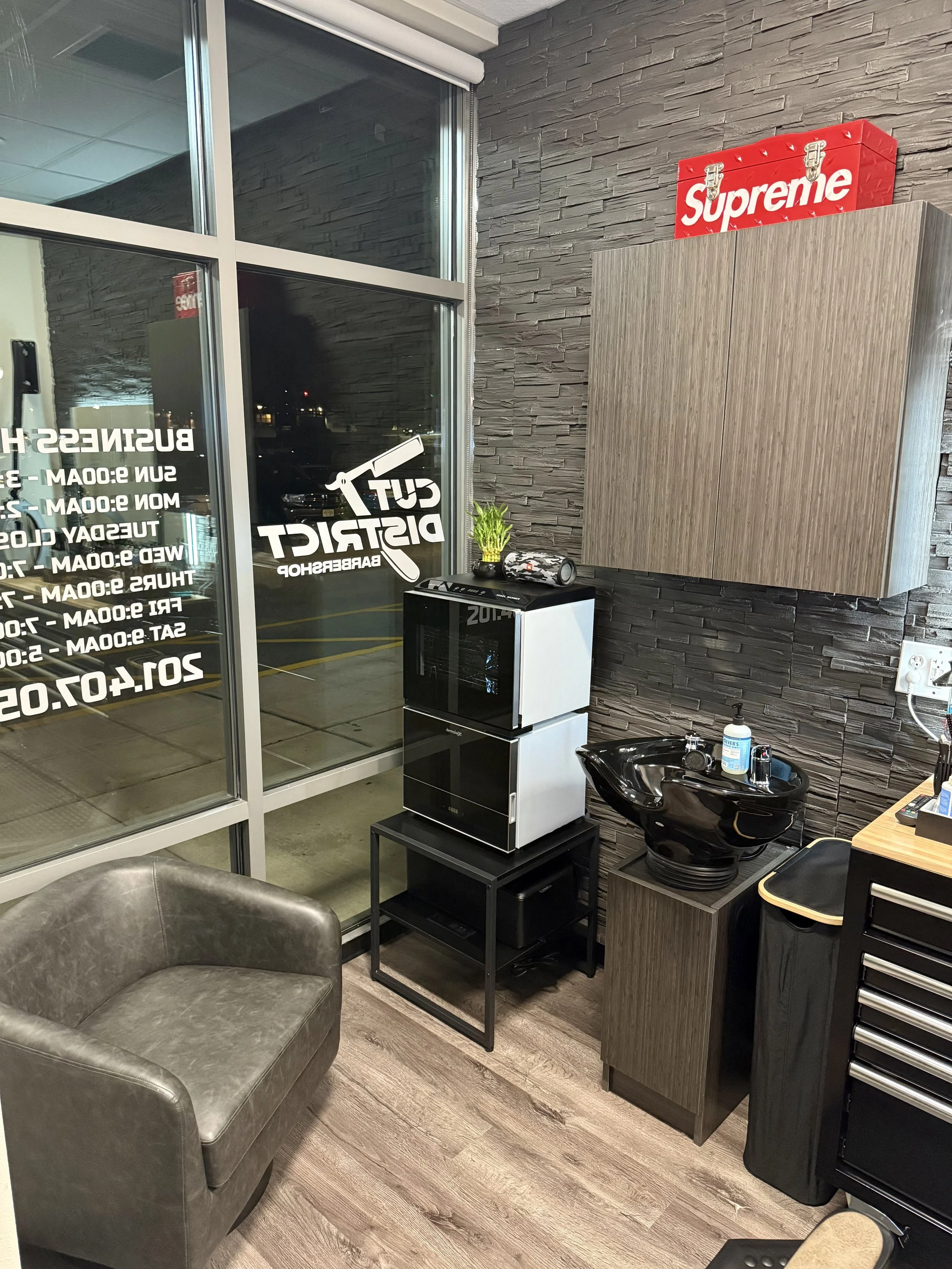 A hair salon or barber shop interior showing a waiting area chair, behind it a window with business hours signage, and a styling station with a black ceramic washbasin, a wooden cabinet, a small black refrigerator, and various equipment.