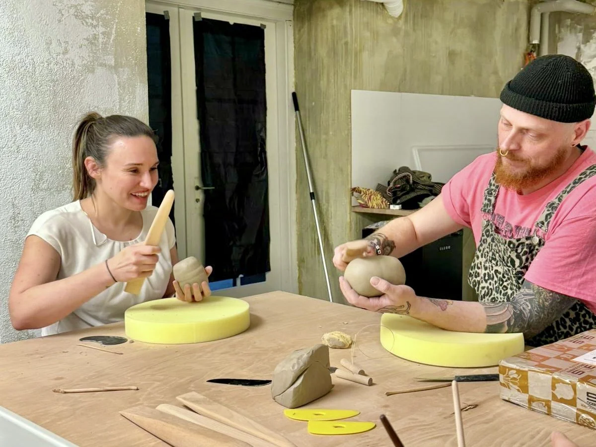 Zwei Menschen, ein Mann und eine Frau, sitzen an einem Tisch und formen Ton. Die Frau lacht und hält ein Werkzeug und eine Tonkugel, während der Mann mit einem Werkzeug an einer Tonkugel arbeitet. Der Tisch hat verschiedene Werkzeuge, Ton und andere 