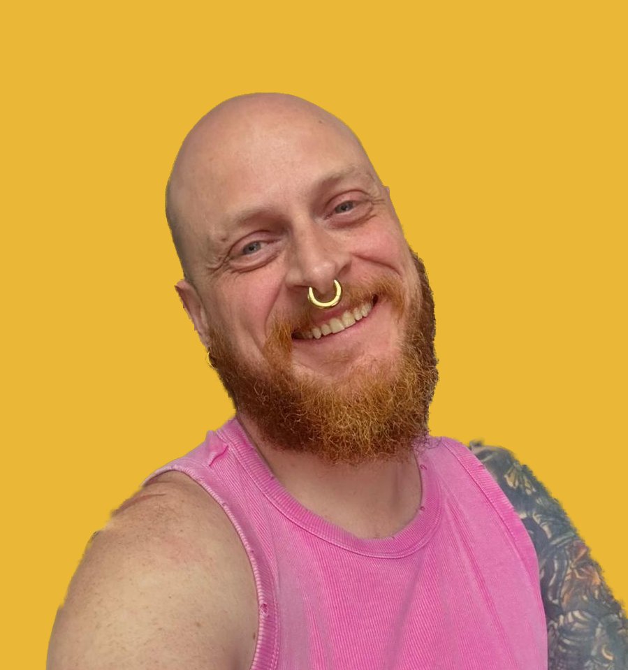 Eine lachende Person mit einem Piercing in der Nase, einem roten Bart, einem rosa Shirt, einer Tätowierung auf dem Arm vor einem gelben Hintergrund.