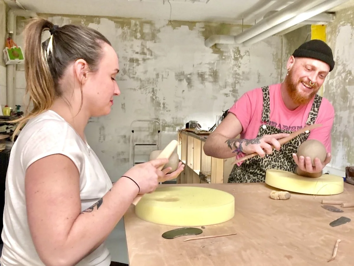 Zwei Menschen in einer Werkstatt, die mit Töpfern beschäftigt sind, eine Person und eine Frau sitzen an einem Holztisch, Die Person lächelt und zeigt an einer Keramikskulptur eine Töpfertechnik, während die Frau an Ihrer Skluptur arbeitet.
