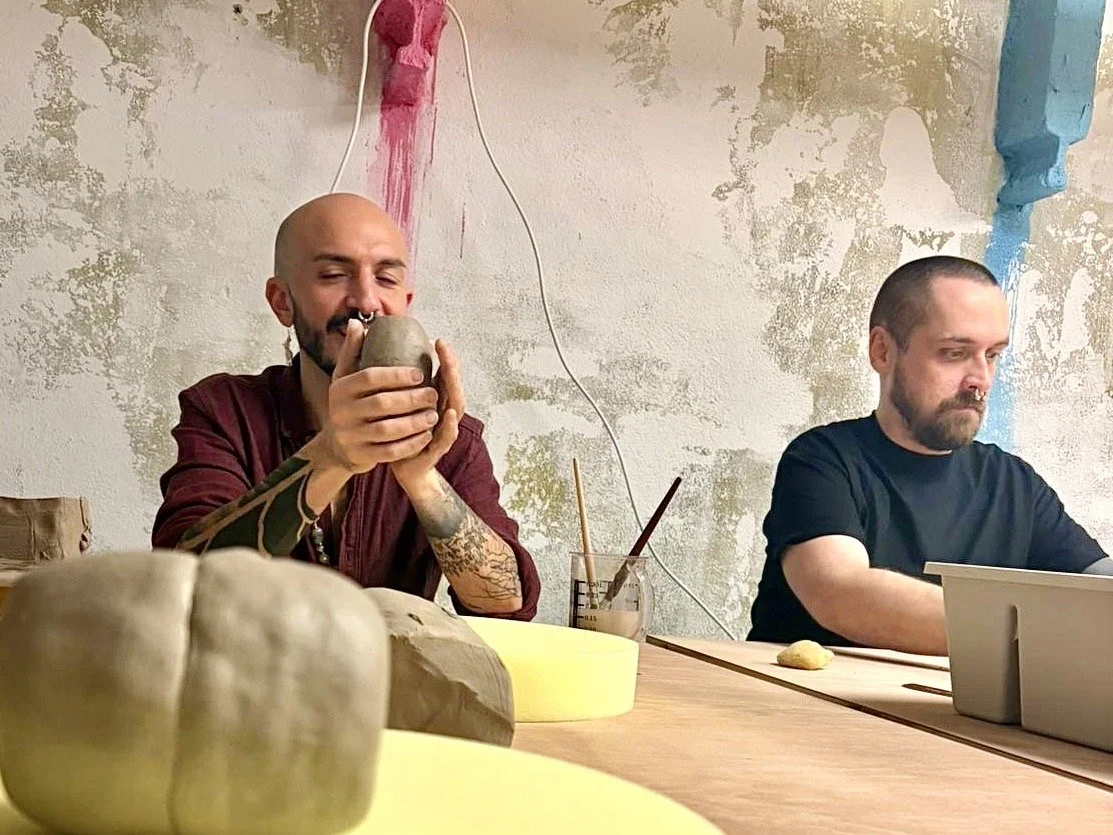 Zwei Personen sitzen an einem Tisch. Die eine Person auf der linken Seite hält eine ungewöhnliche, handgefertigte Vase und scheint sie zu bewundern, während Die andere Person auf der rechten Seite auf konzentriert arbeitet.