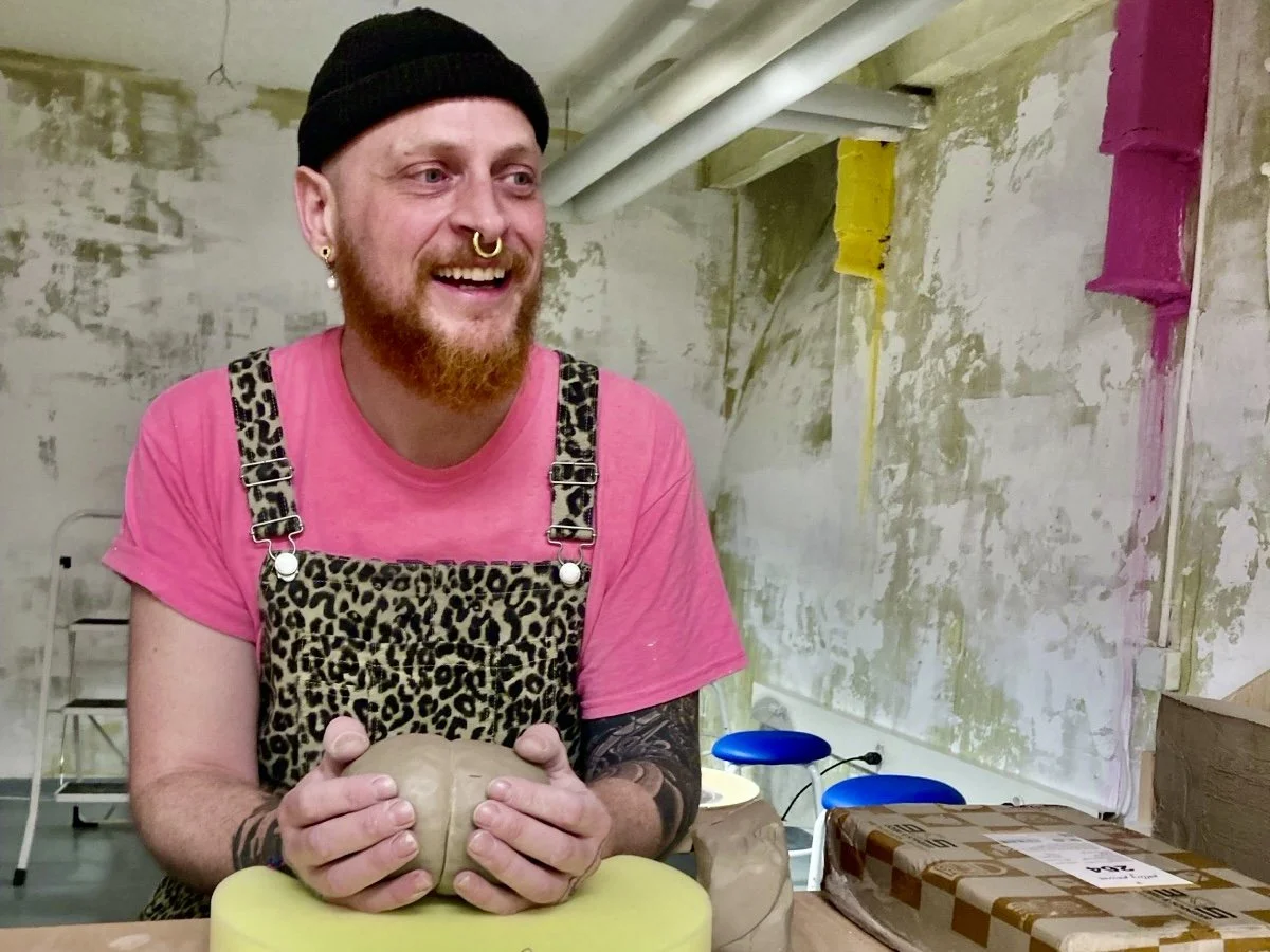 Ein lachender junger Mann mit rotem Bart, Piercings und einem schwarzen Hut hält eine Tonfigur in den Händen. Er trägt ein pinkes T-Shirt und ein Leo-Print-Schürze. Die Szene scheint in einem Kunst- oder Handwerksraum stattzufinden.
