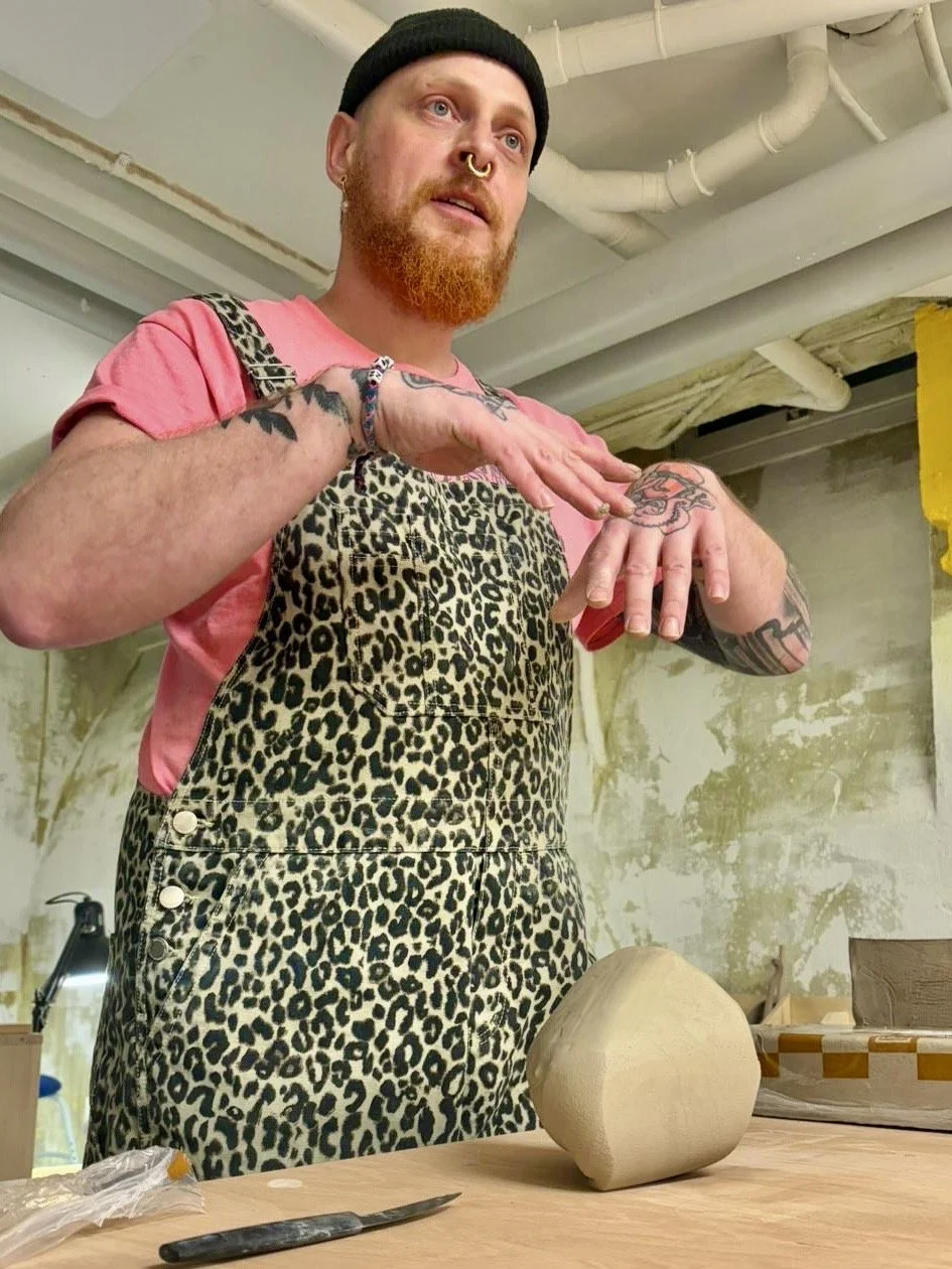 Eine Person mit roten Haaren, Bart, Piercings und Tattoos arbeitet in einer Werkstatt an einem Töpferprojekt. Sie trägt eine schwarze Mütze, eine rosa T-Shirt und einen Leopard Print. Sie steht an einem Tisch mit einer Tonfigur und Werkzeug.