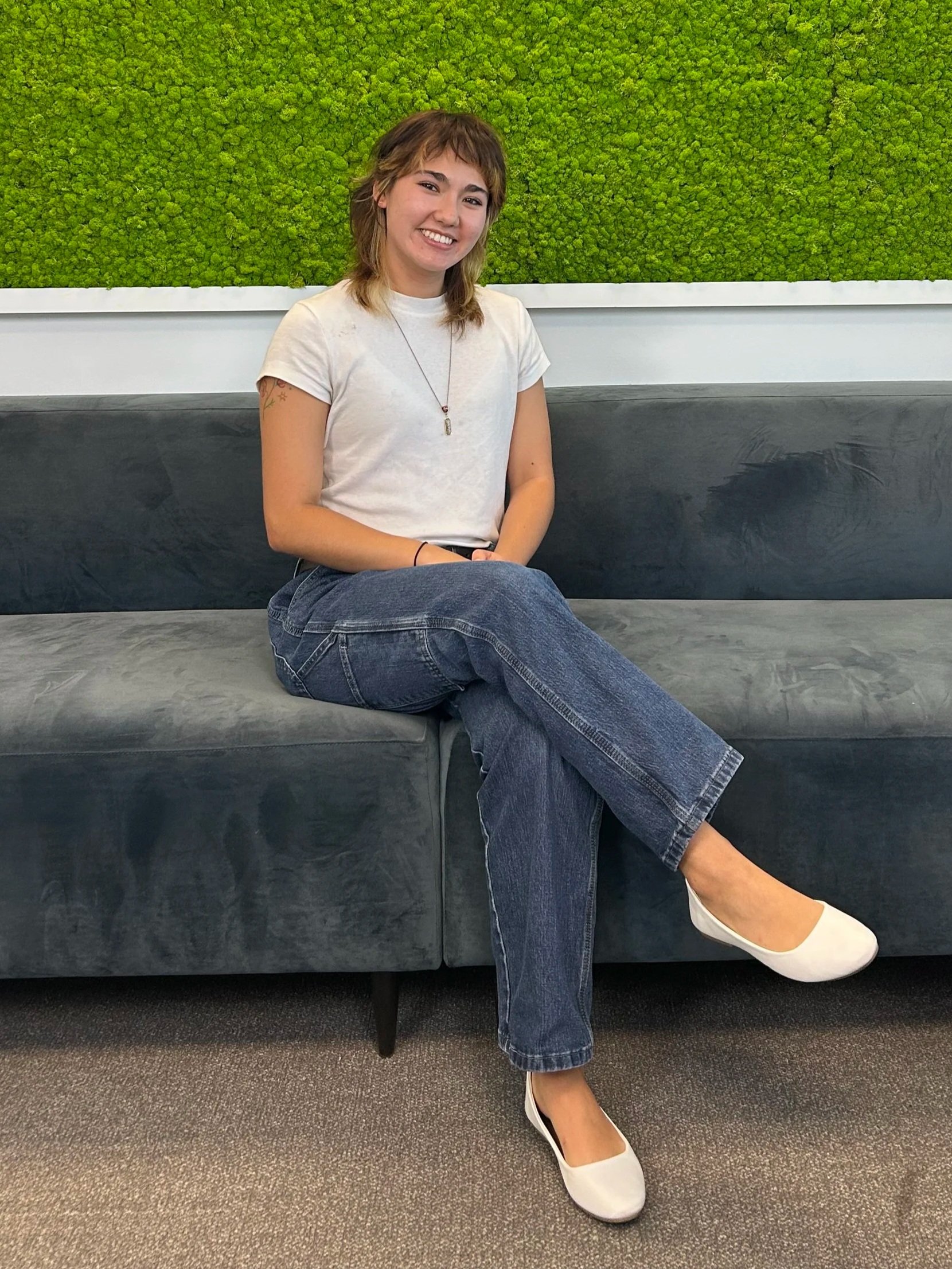 A young woman with shoulder-length brown hair, dressed in a white t-shirt and jeans, sitting on a dark gray sofa in front of a green moss wall, smiling.