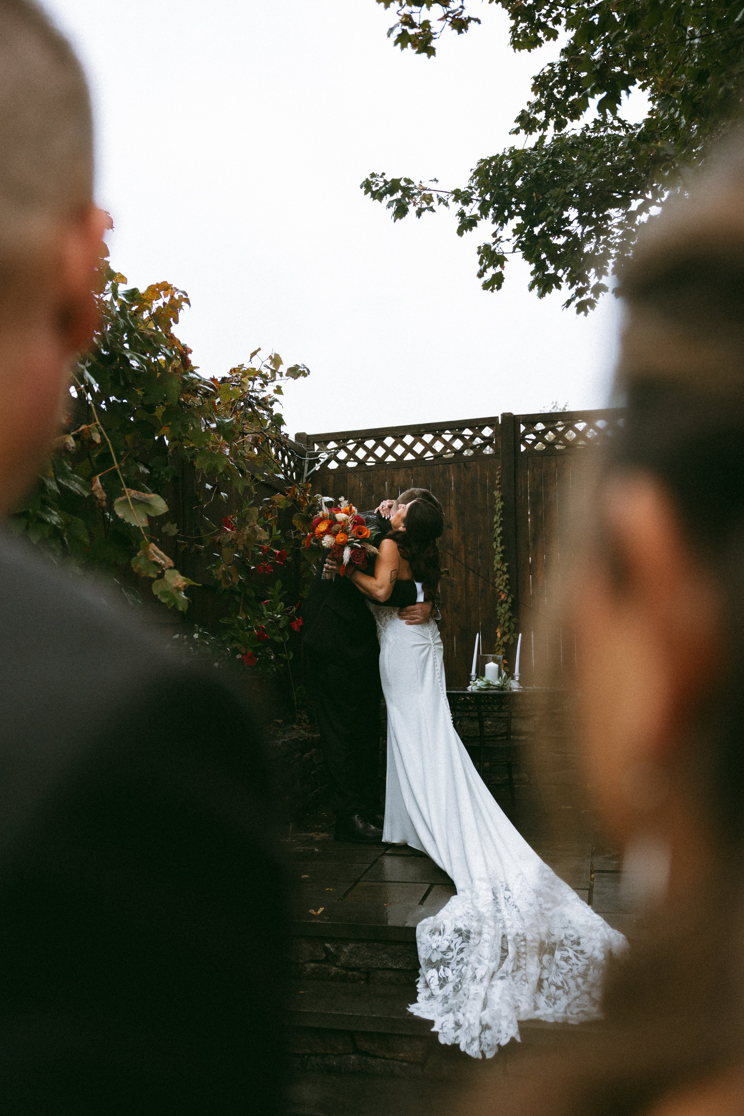 Couple kissing at a wedding outdoors, with blurred guests in foreground, a wooden fence, greenery, and a small table with candles in the background.