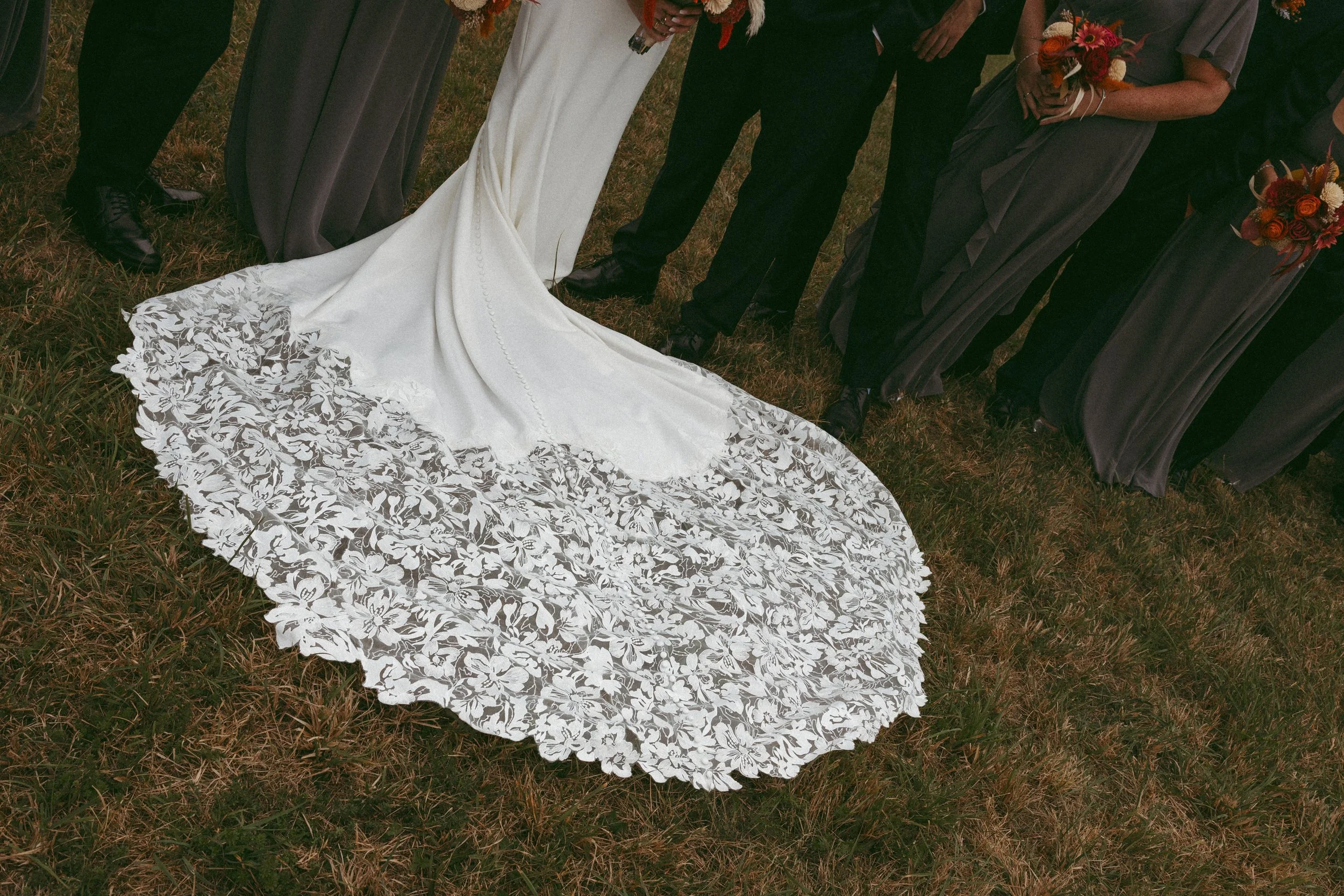 Wedding party standing outdoors on grass, centered around a bride in a white gown with an intricate lace train, surrounded by bridesmaids in gray dresses holding bouquets, and groomsmen in suits.