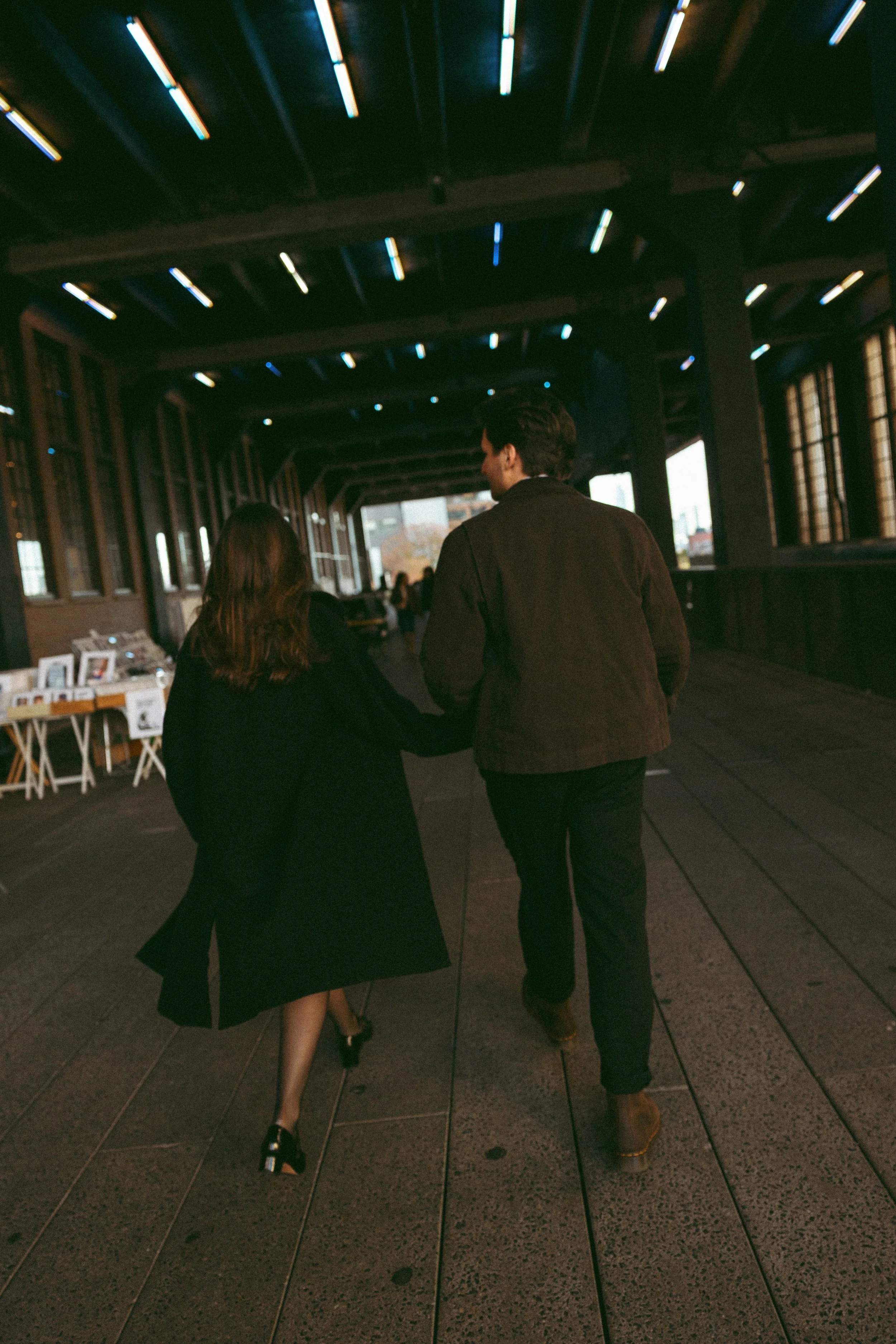 A man and woman walking hand in hand in a covered outdoor walkway with modern lighting on the ceiling.