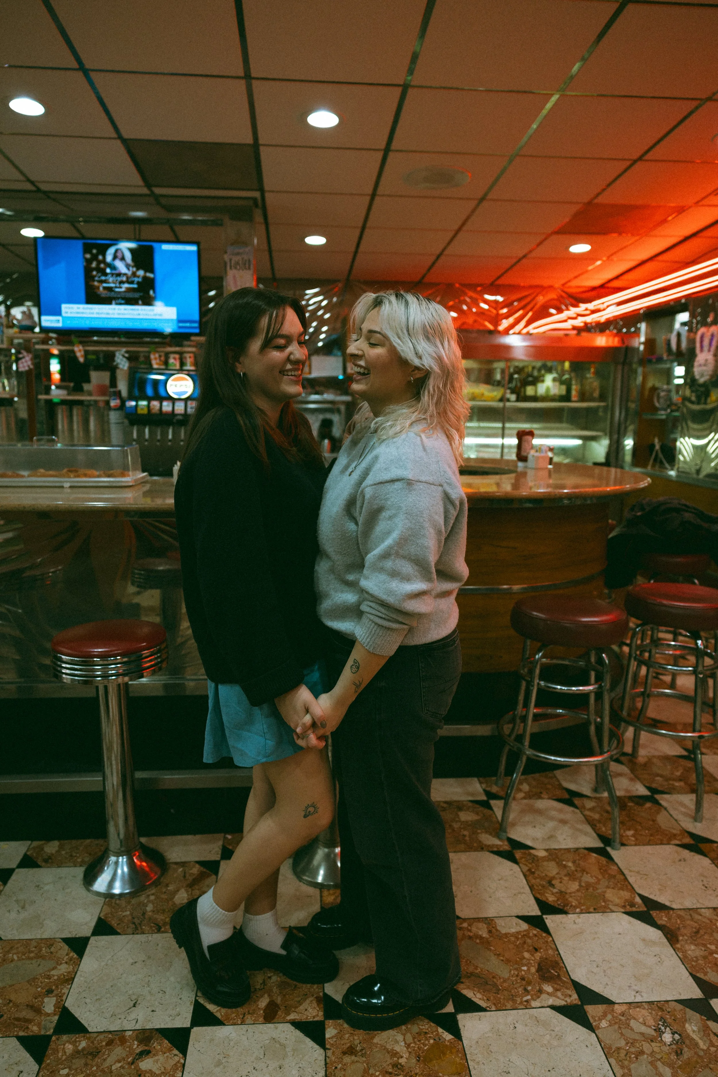 Two women holding hands and smiling at each other in a retro-style diner with checkered tile floor and bar stools.