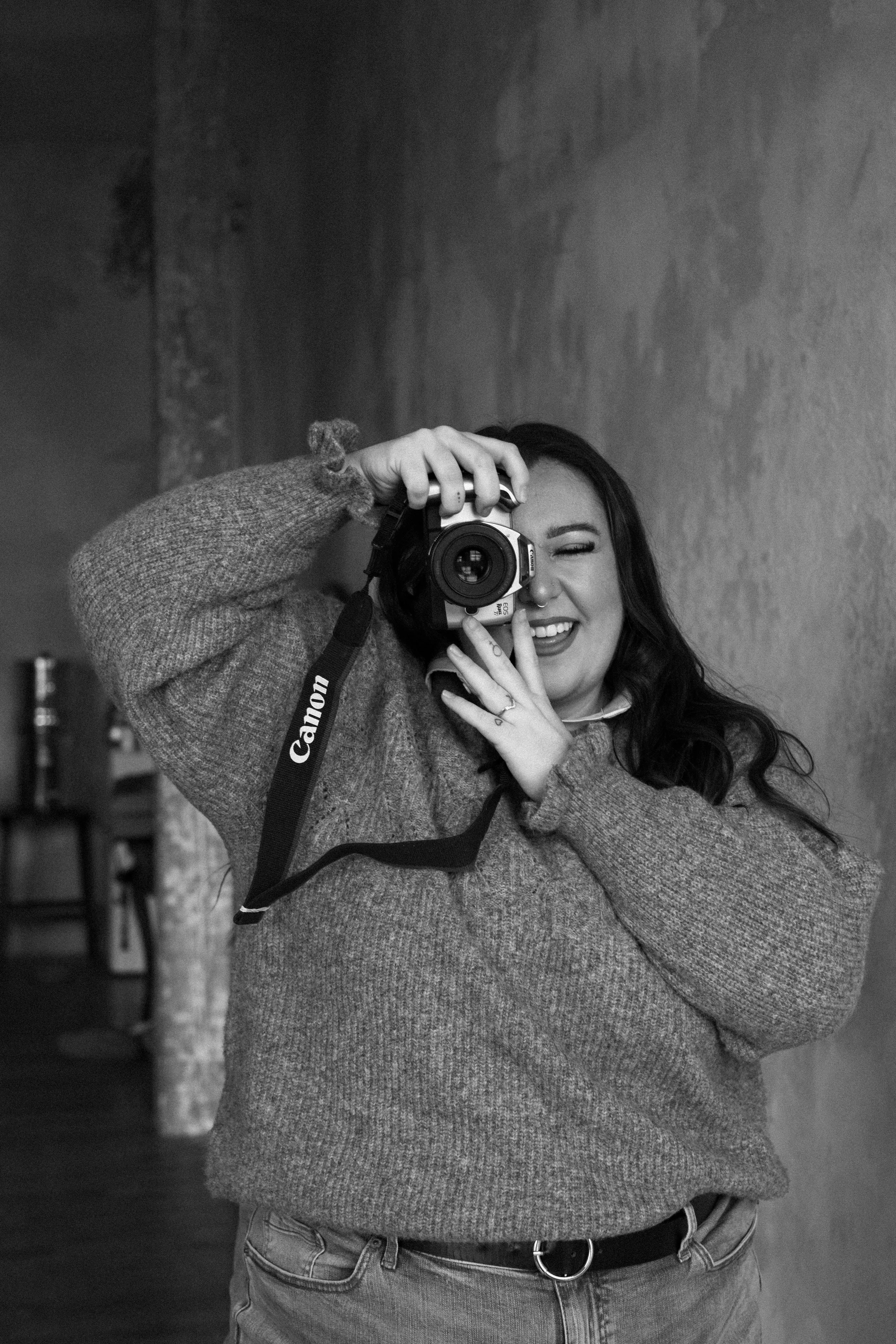 A woman taking a photograph with a Canon camera, smiling and holding the camera up to her face while winking. She has long dark hair and is wearing a cozy sweater.